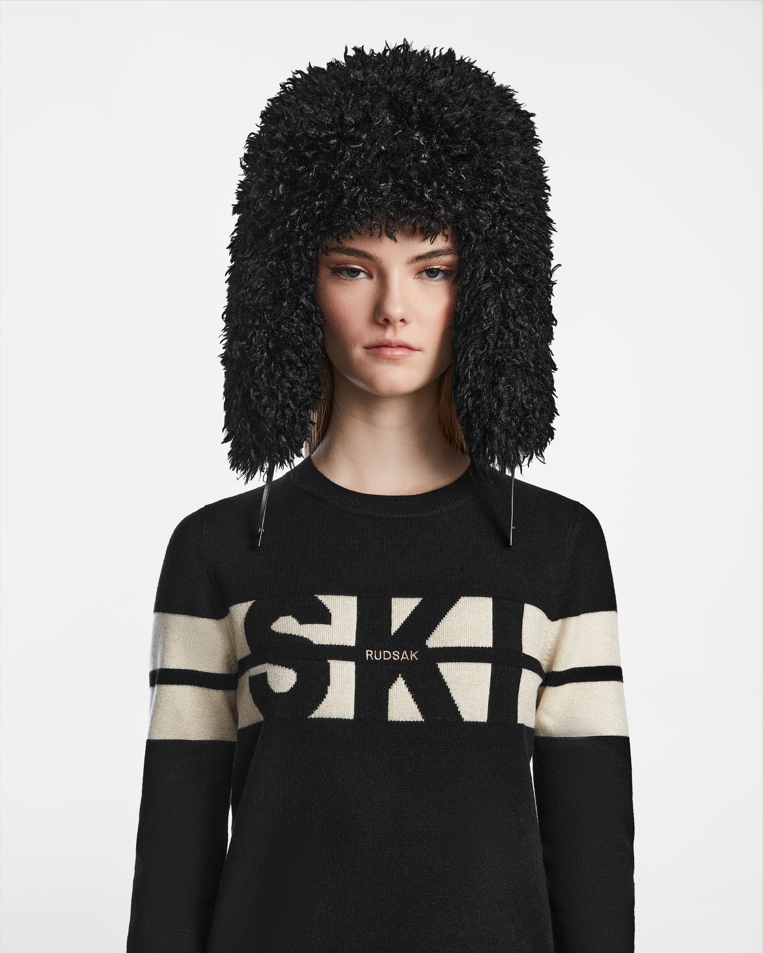 A woman in a Rudsak DARBY M - 9424510 Black faux Mongolian fur trapper hat and a black SKI RUDSAK sweater with white stripes, standing against a plain white background—a cozy, chic winter look.