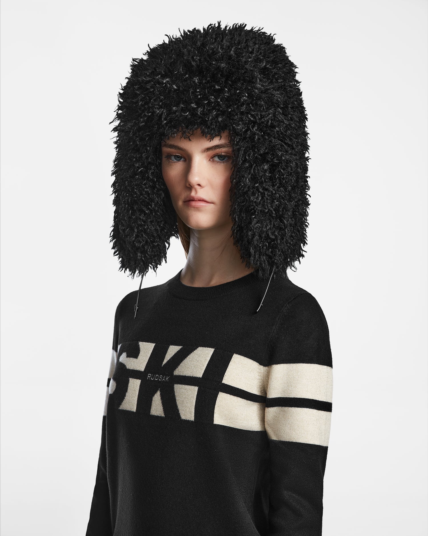 A woman wears the Rudsak DARBY M - 9424510 Black faux Mongolian fur unisex aviator hat with ear flaps and a black sweater with white stripes, standing against a white background, looking at the camera with a neutral expression.