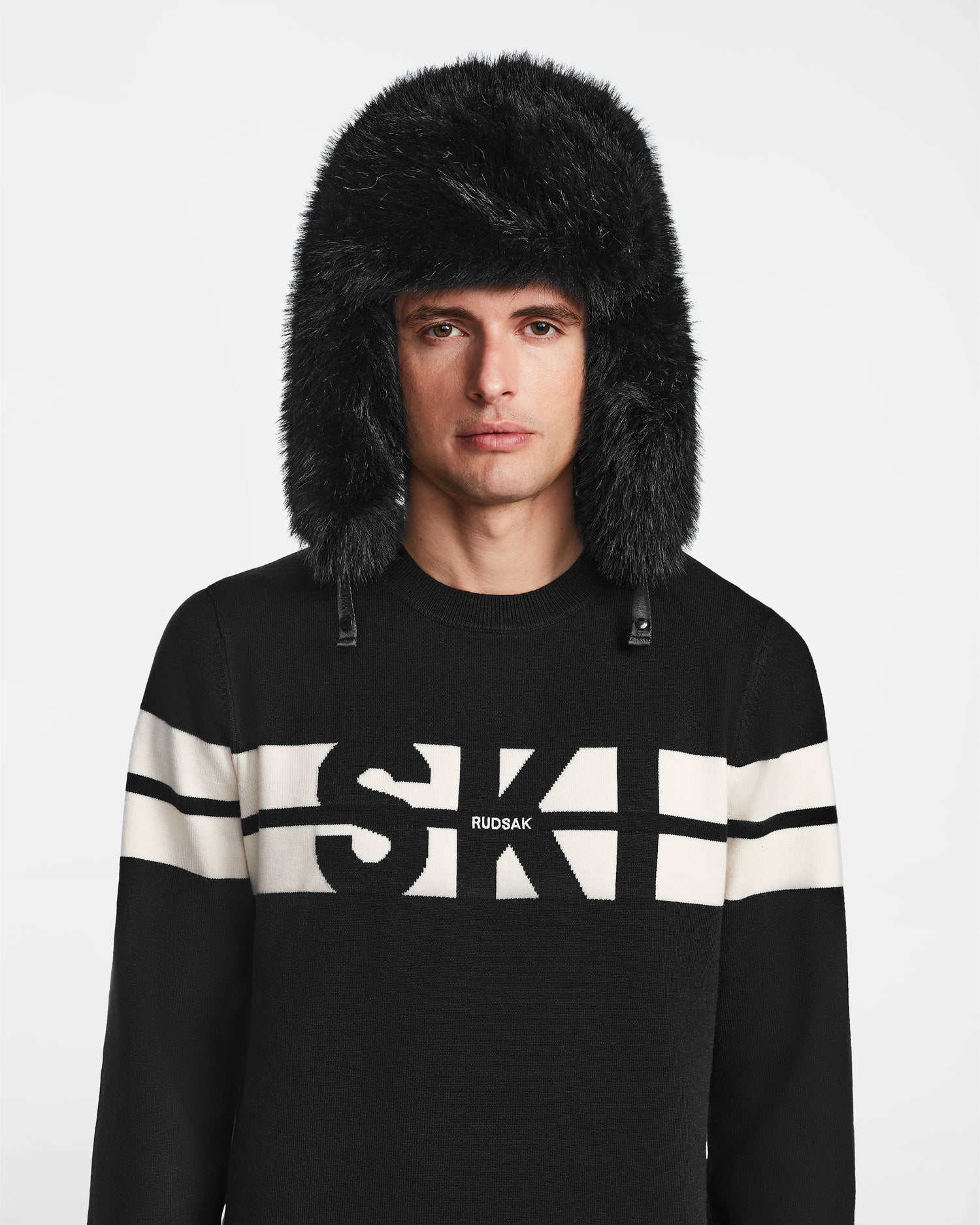 A man in a black sweater with white stripes and SKI RUDSAK text stands against a white background, wearing the DARBY - 9422510 BLACK unisex aviator hat with faux fur by Rudsak—a classic winter look.