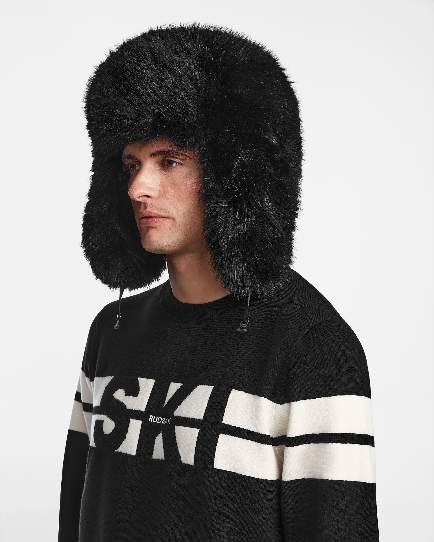 A man wears the Rudsak DARBY - 9422510 BLACK unisex aviator hat with large, fluffy black faux fur ear flaps, paired with a black sweater featuring white stripes and "SKI" text, set against a plain white background.