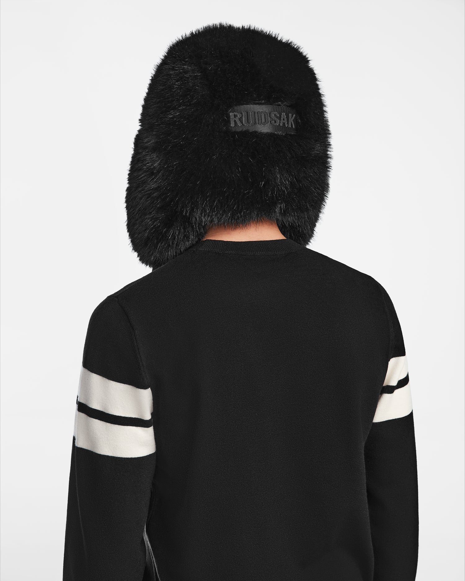 A person wearing the Rudsak DARBY - 9422510 BLACK faux fur winter hat and a black sweater with white striped sleeves, shown from behind against a plain white background.