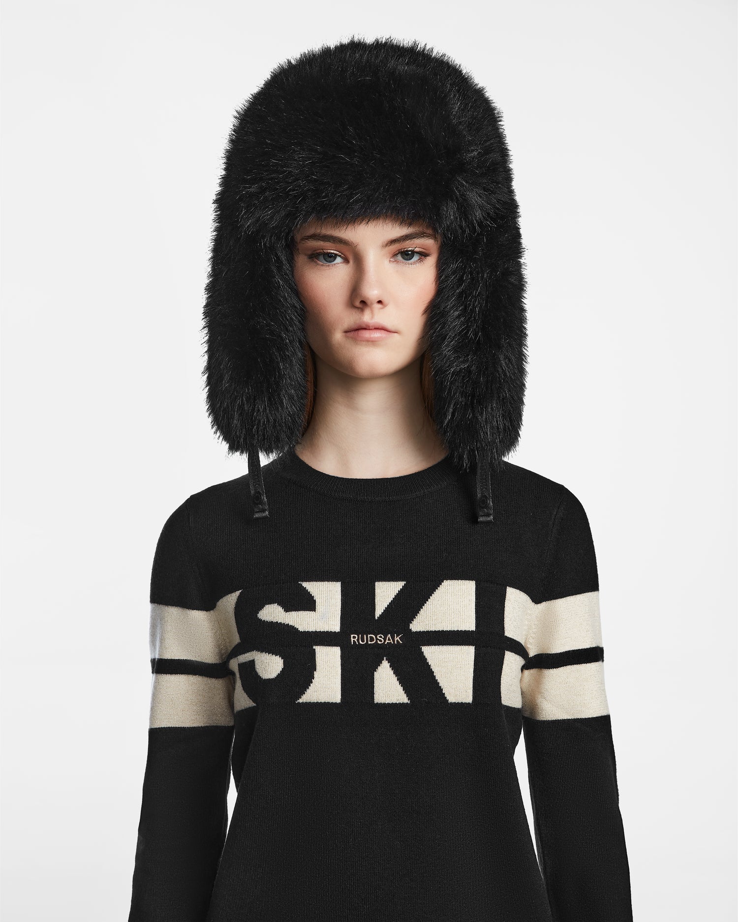 A woman wears the Rudsak DARBY - 9422510 BLACK faux fur winter hat and a black sweater with white "SKI" lettering, posing against a plain white background.