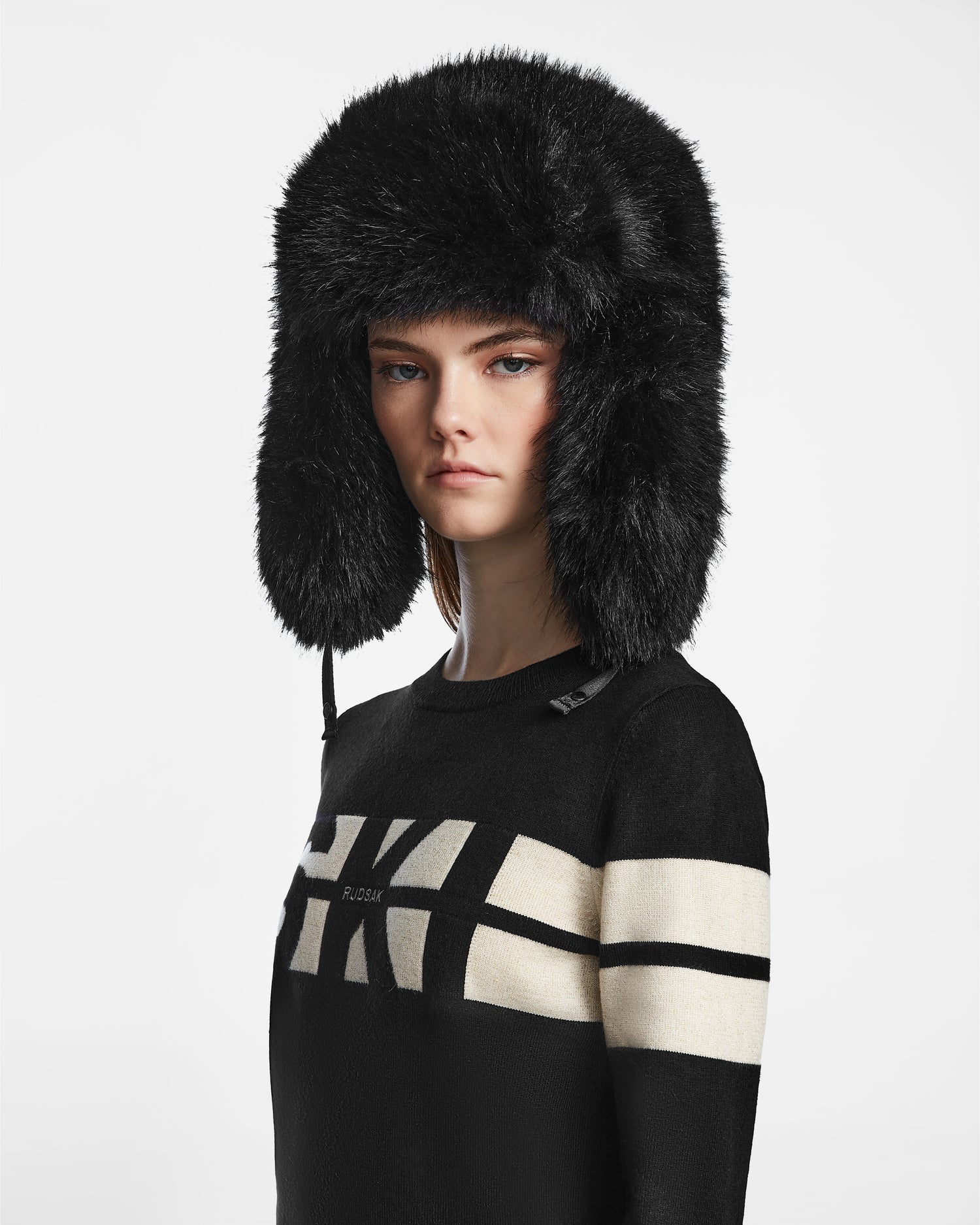 A woman models the Rudsak DARBY - 9422510 BLACK faux fur winter hat with ear flaps and a black sweater with white geometric patterns, standing against a plain light background.