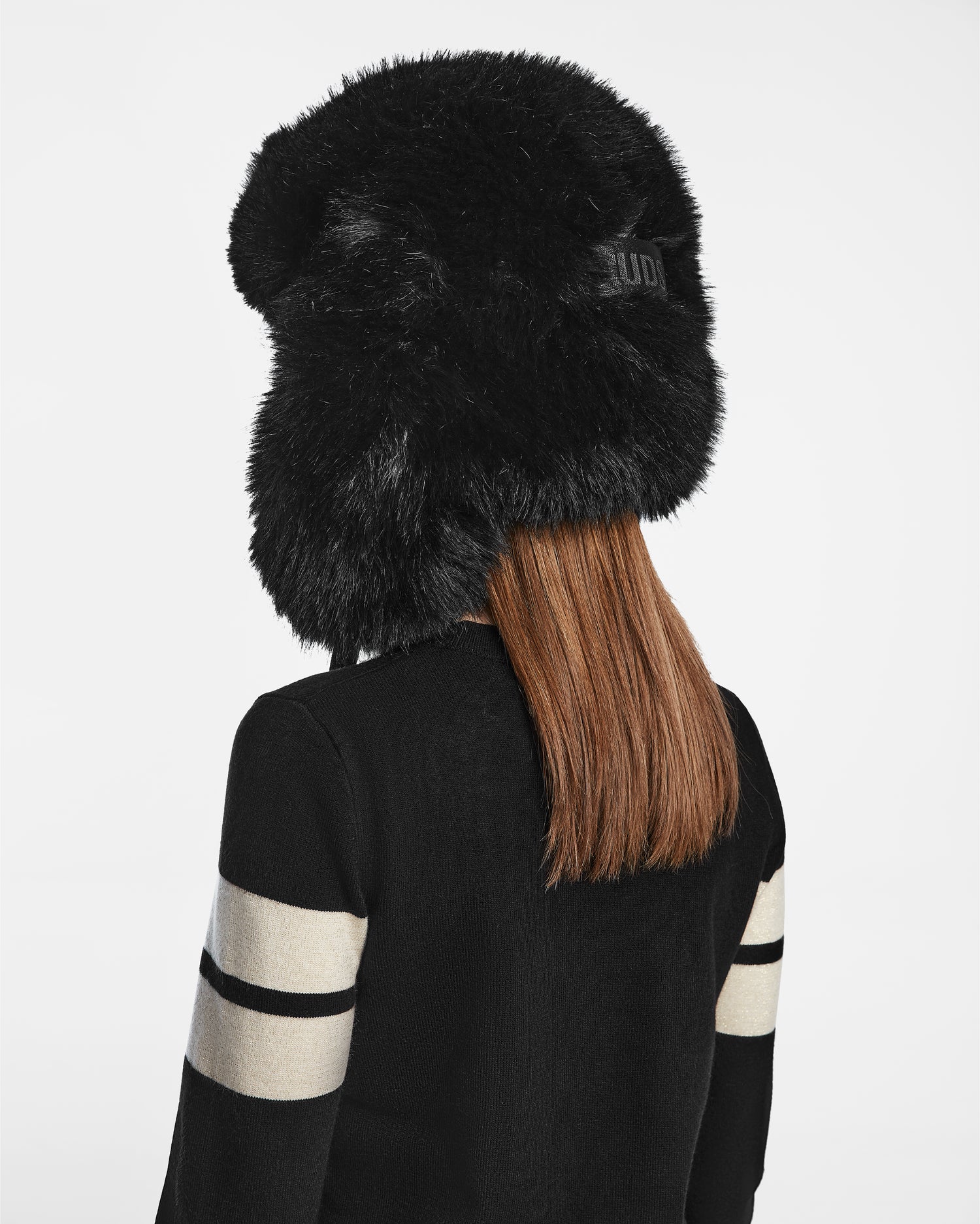 A person with straight, light brown hair is shown from behind wearing the Rudsak DARBY - 9422510 BLACK faux fur winter hat and a black sweater with white striped sleeves, set against a plain white background.