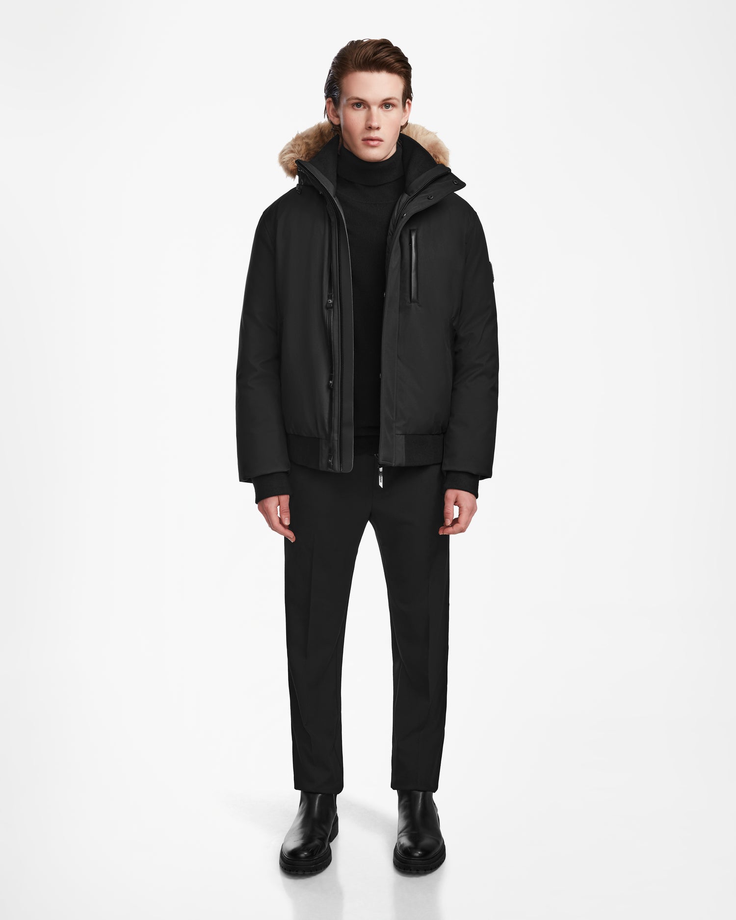 A man faces forward against a white background, wearing the Rudsak DEACON - M123513 Black/Natural bomber-style down puffer with recycled leather trim and RDS-certified down, paired with a black turtleneck, pants, and boots. His hands rest at his sides.