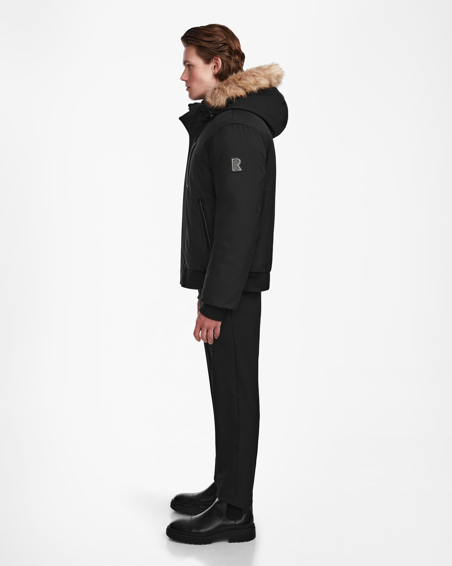 A person stands in profile facing left, wearing the Rudsak DEACON - M123513 Black/Natural down puffer jacket with fur trim, paired with black pants and boots against a plain white background.