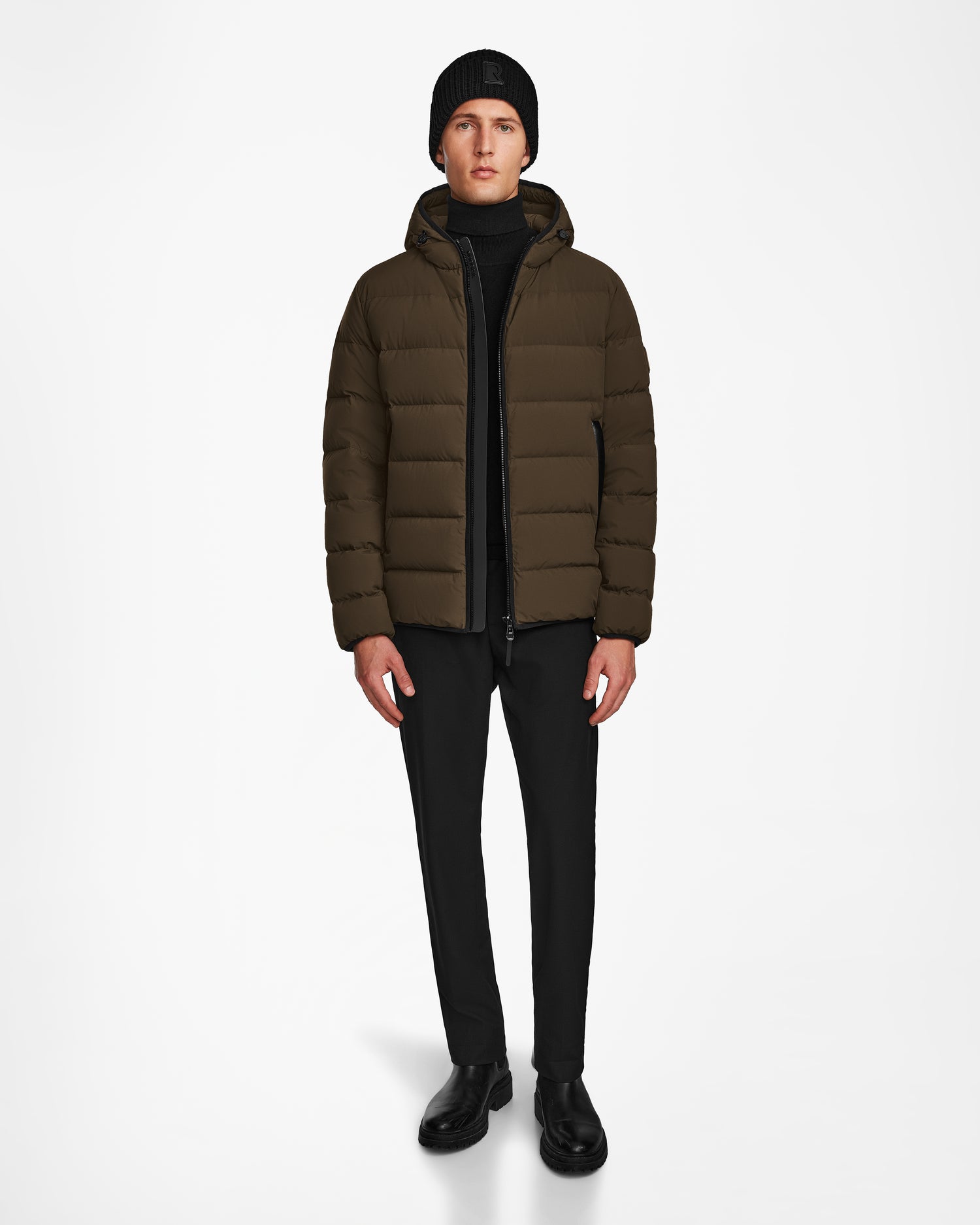 A person stands against a plain white background wearing the Rudsak DEMIAN M - M123507 Army men’s down puffer jacket, made from sustainable recycled fabric, paired with black pants, boots, sweater, and a knit beanie.