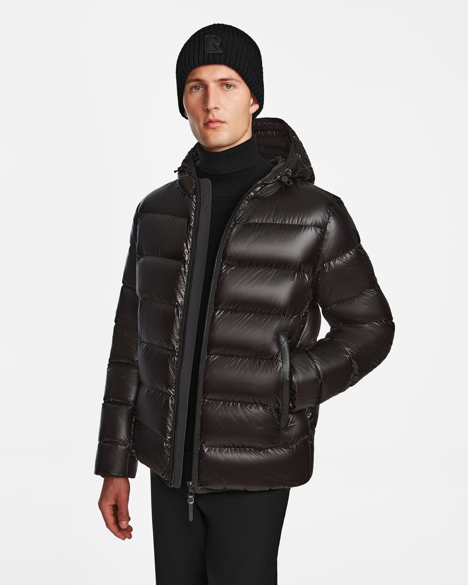 A man stands against a white background wearing the Rudsak DEMIAN S - M123508 Black down puffer jacket, paired with a black knit beanie and black pants, with one hand in his pocket.