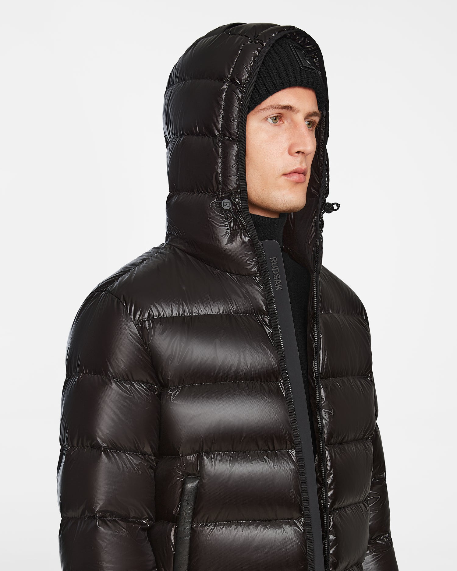 A person wearing the Rudsak DEMIAN S - M123508 Black mens down puffer jacket with the hood up and a black knit beanie, looking to the side against a plain white background.