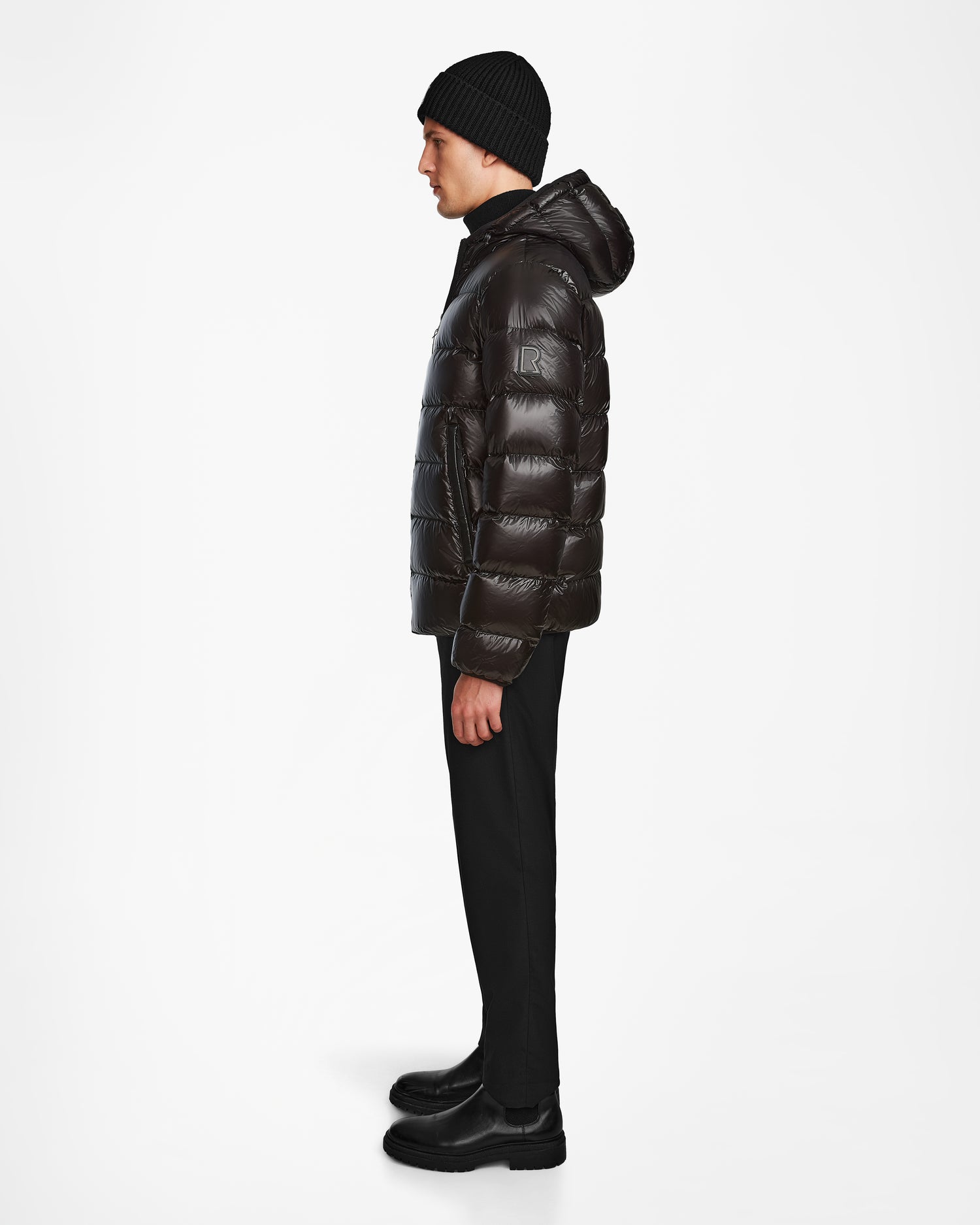 A man stands in profile against a white background, wearing the Rudsak DEMIAN S - M123508 Black men’s down puffer jacket, paired with black pants, black boots, and a black knit beanie.