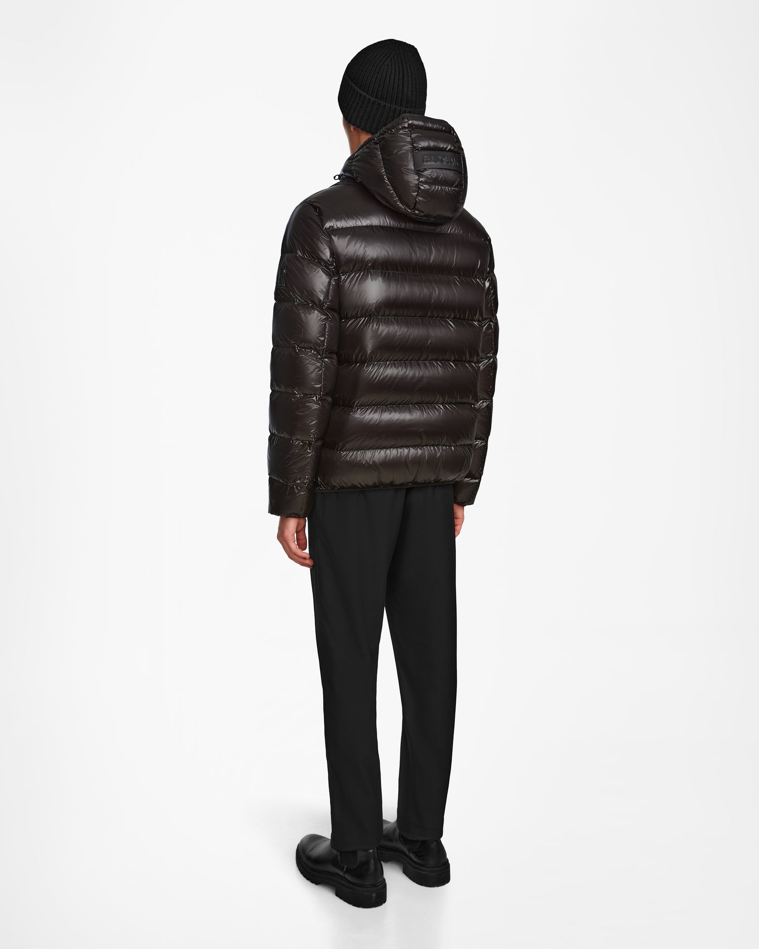 A person stands facing away in a Rudsak DEMIAN S - M123508 Black mens shiny down puffer jacket with hood, water- and wind-resistant, paired with black pants, shoes, and a knit beanie against a plain white background.