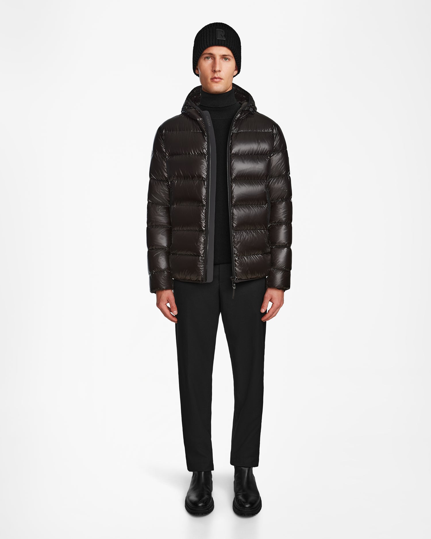 A man faces forward wearing the Rudsak DEMIAN S - M123508 Black mens down puffer, paired with a black beanie, black pants, and boots against a plain white background.