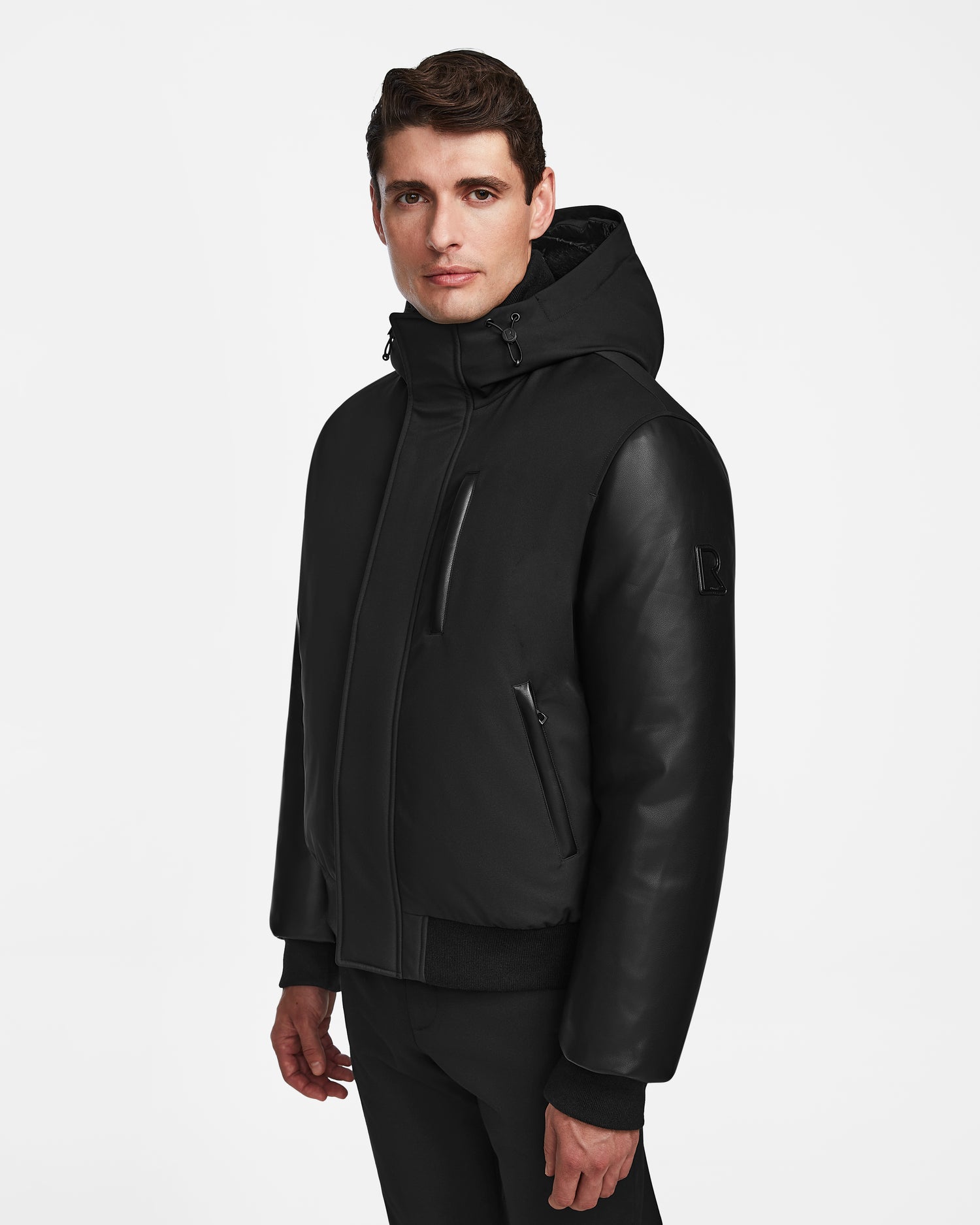 A man with short brown hair wears the Rudsak DERON - M123517 BLACK mens bomber jacket, featuring zippered pockets and weather-resistant material, standing against a plain white background.