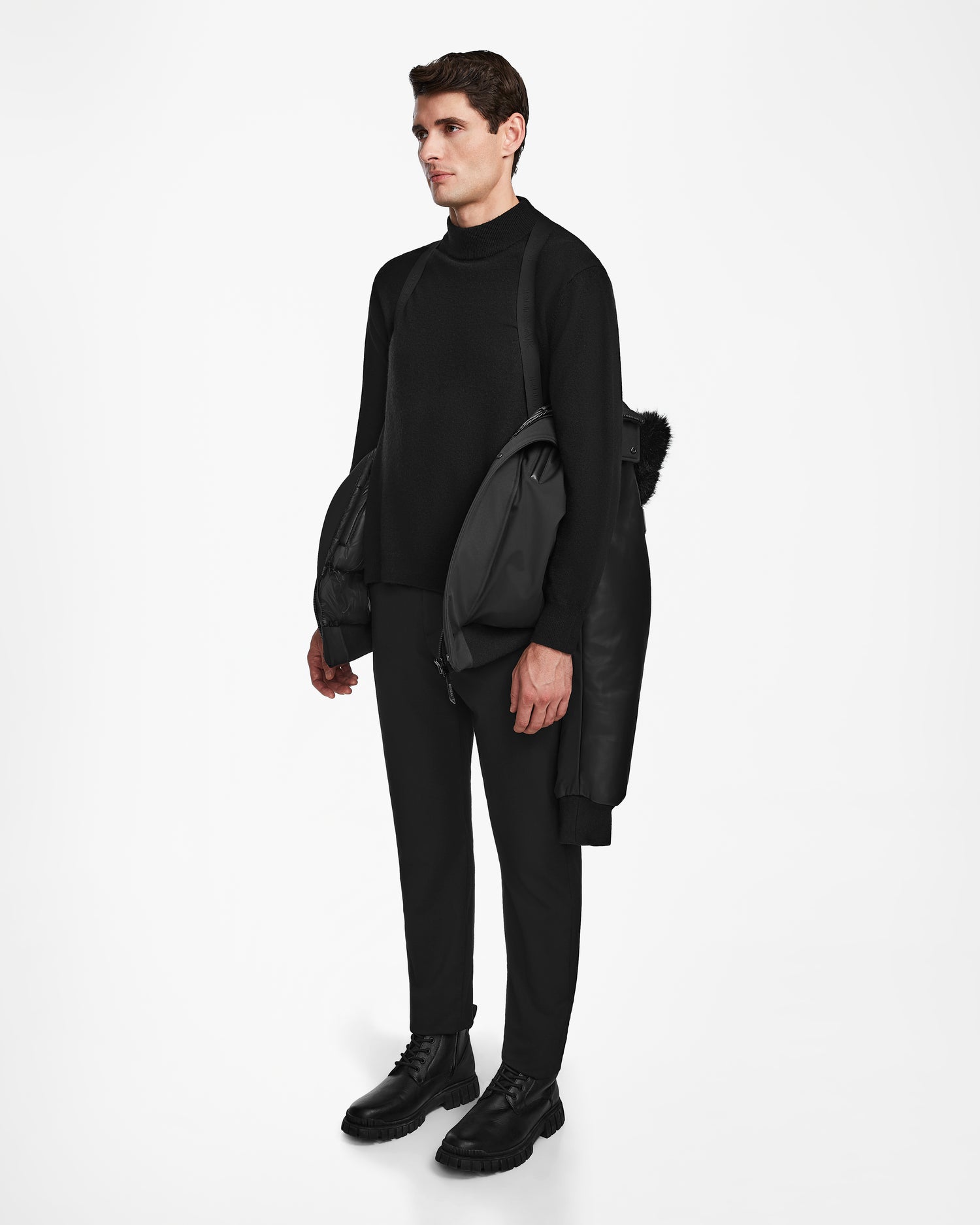 A man stands against a white background in black attire, sporting the weather-resistant Rudsak DERON - M123517 BLACK men’s bomber jacket draped over his shoulders. He gazes to the side, effortlessly blending style and function.