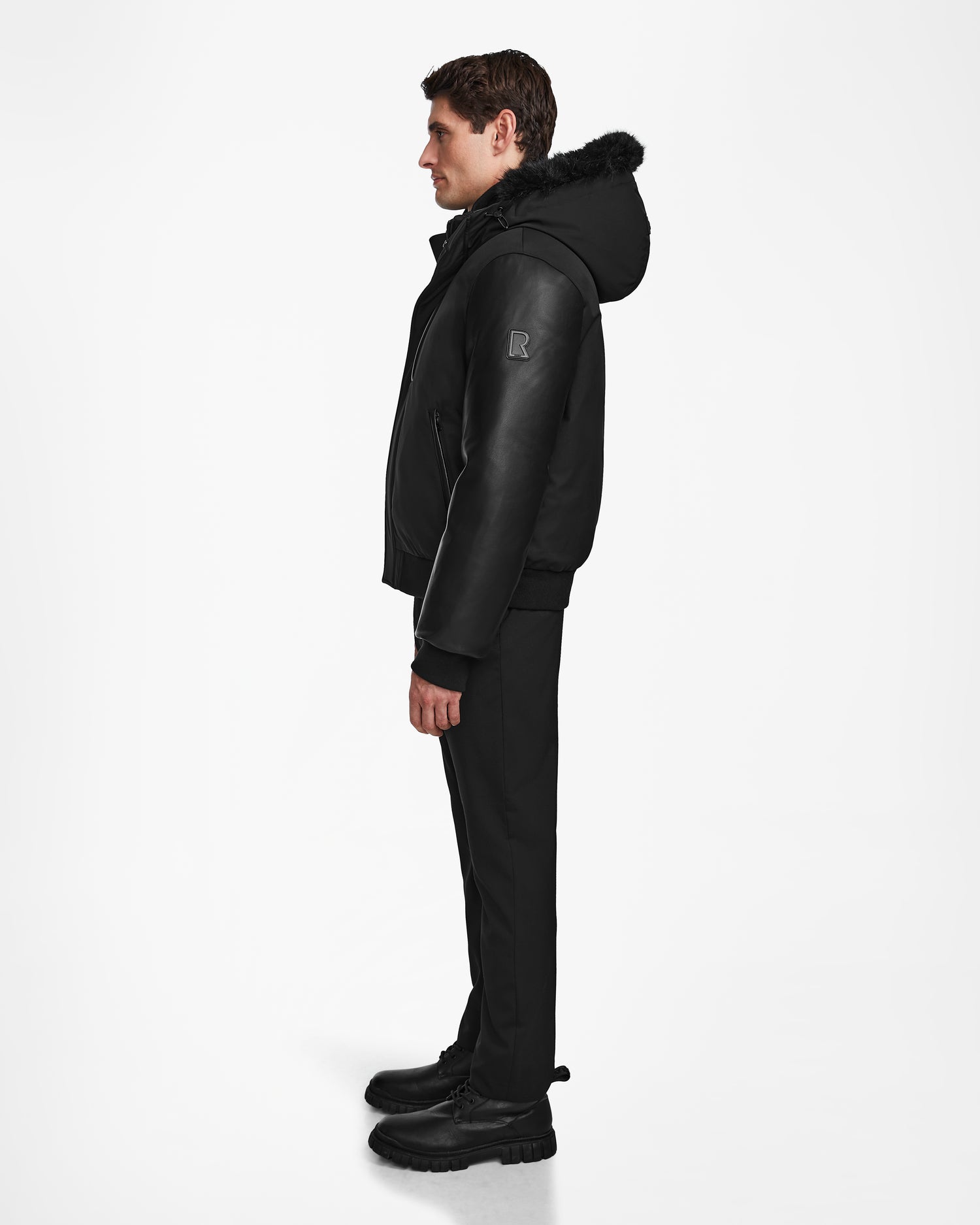 A man stands in profile wearing the Rudsak DERON - M123517 BLACK weather-resistant mens bomber jacket, paired with black pants and boots, against a plain white background.