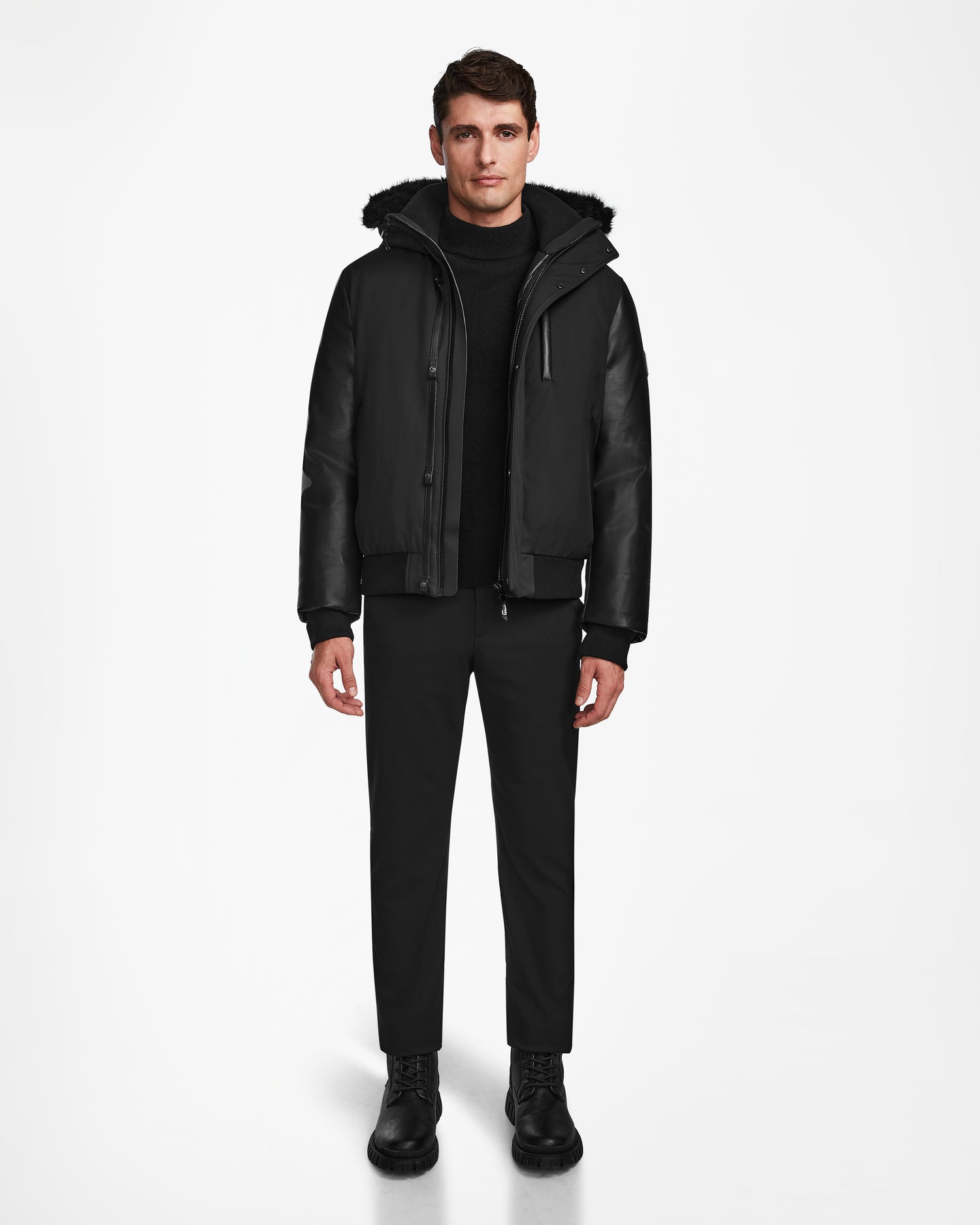 A man stands facing forward in the Rudsak DERON - M123517 BLACK mens bomber jacket, featuring leather sleeves and weather resistance, paired with black pants, sweater, and boots against a plain white background.