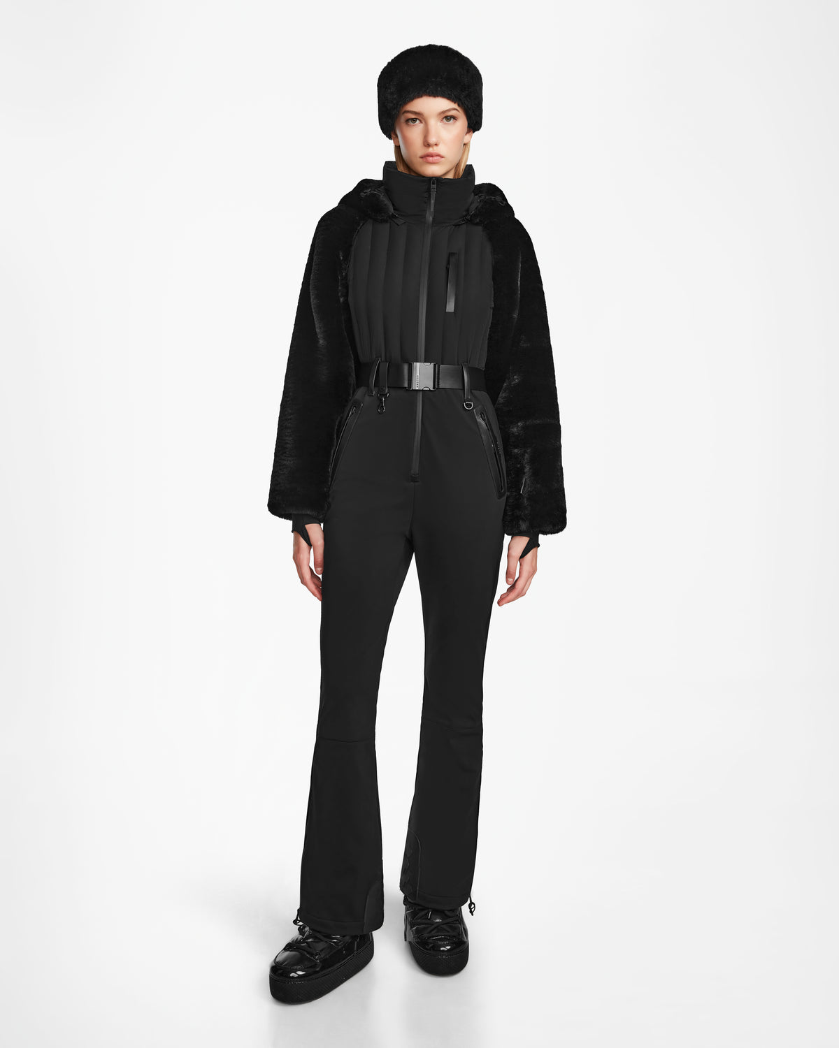 A person stands facing forward in the Rudsak DESTINY - 8124727 Black one-piece ski suit with RECCO rescue technology, paired with a black fur hat and shoes, against a plain white background.