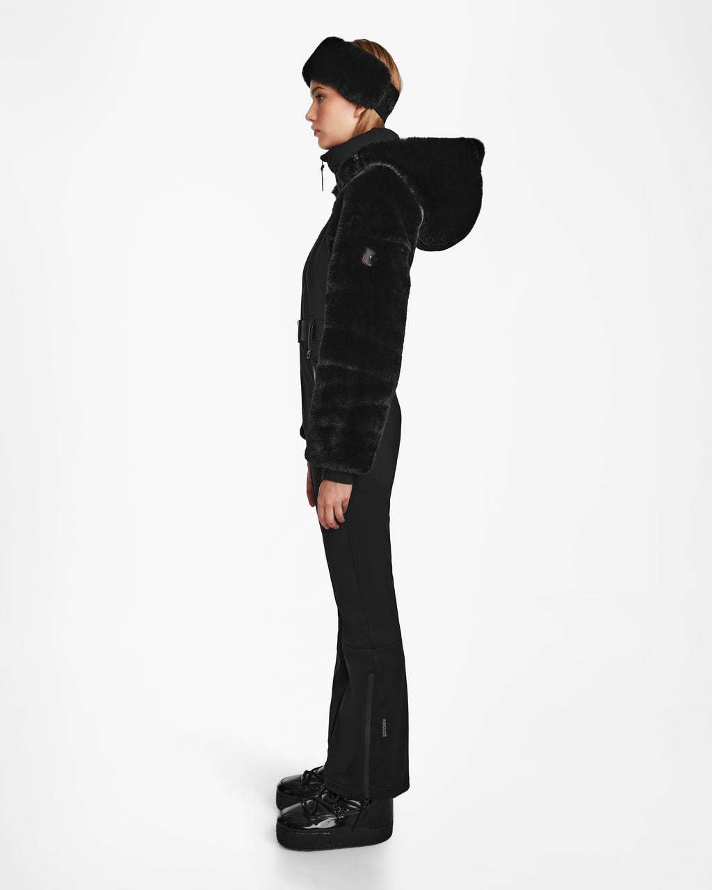 A person in profile models the Rudsak DESTINY - 8124727 Black womens ski suit with RECCO rescue tech, a fur-lined hood, a wide headband, and matching shoes, set against a plain white background.