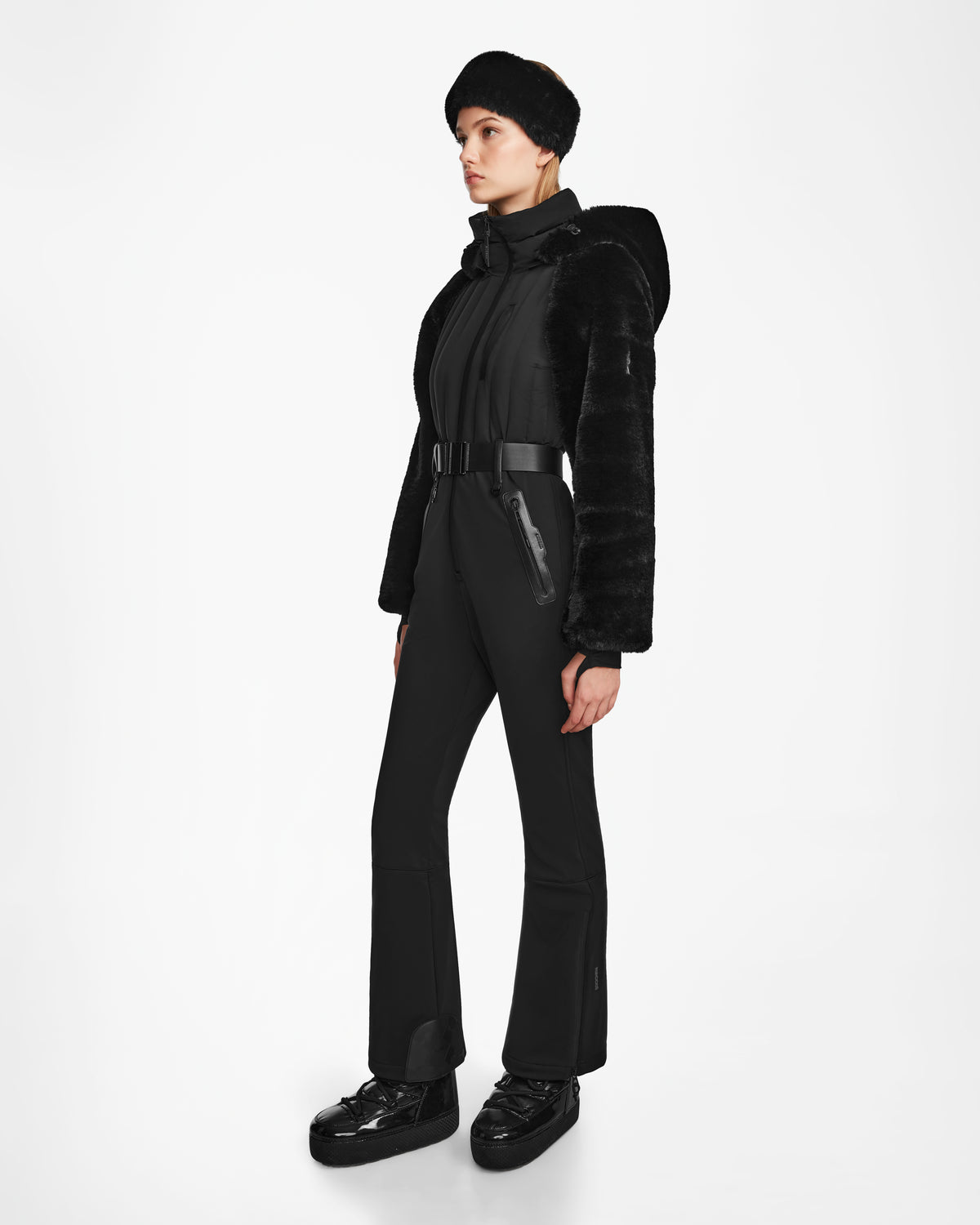 A woman stands against a white background wearing the Rudsak DESTINY - 8124727 Black one-piece ski suit with fur sleeves and platform shoes, looking to the side. The modern outfit features advanced RECCO rescue technology.