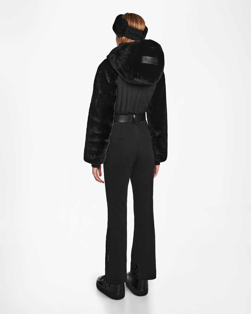 A woman faces away, wearing the Rudsak DESTINY - 8124727 Black faux fur jacket with a high collar over a sleek ski suit, black sneakers, and a black headband, set against a plain white background.