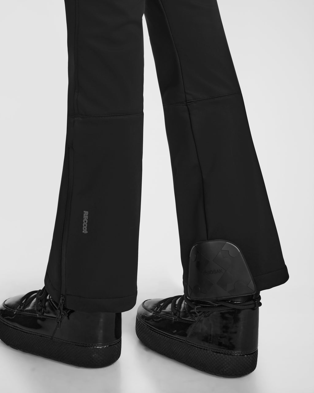 A close-up of a person wearing Rudsak DESTINY - 8124727 Black flared pants with RECCO rescue tech, glossy black shoes, and a black rectangular device above the right shoe against a light background.