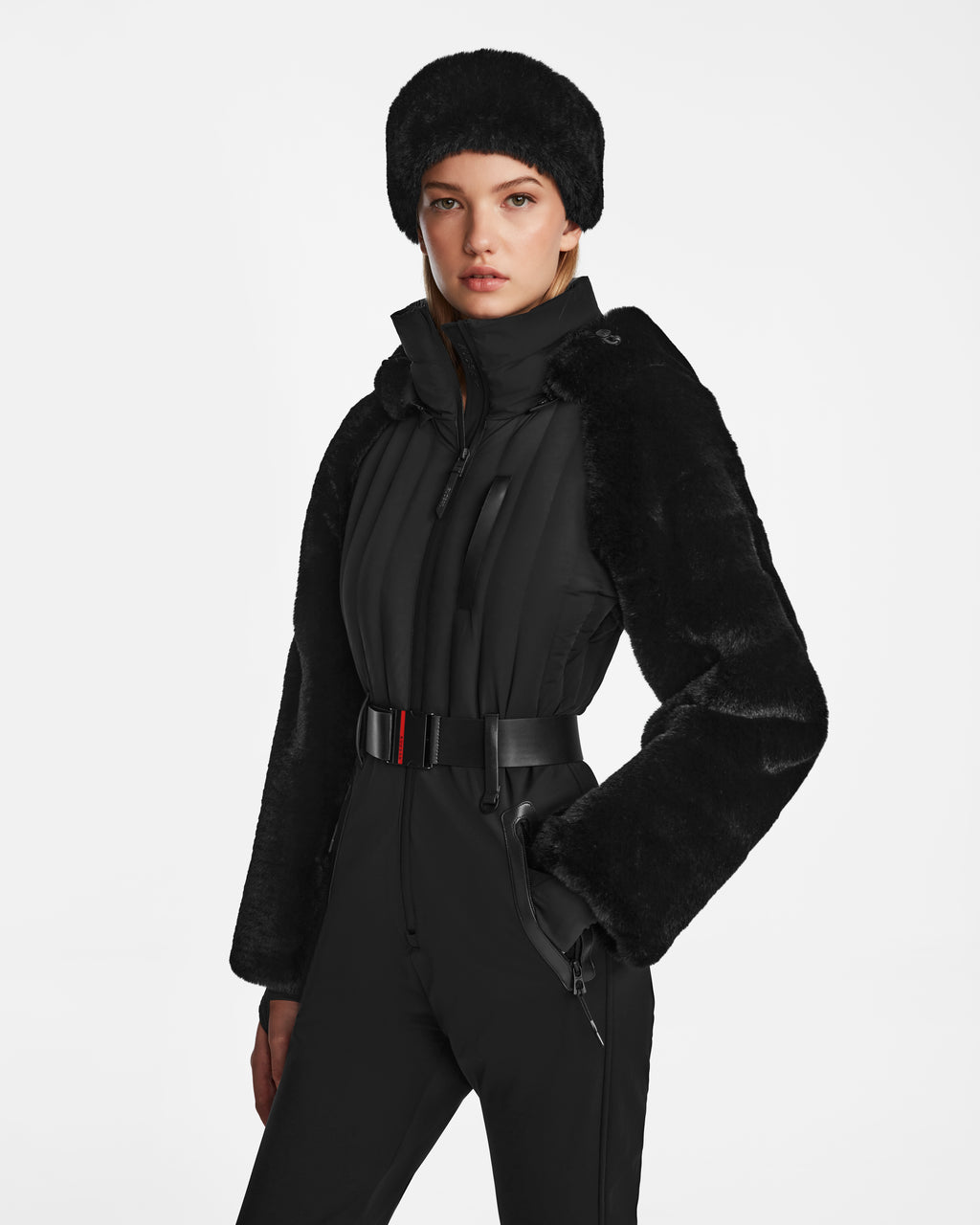 A woman stands against a plain white background, wearing the Rudsak DESTINY - 8124727 Black one-piece ski suit with a belt, faux fur sleeves, and a matching faux fur hat.