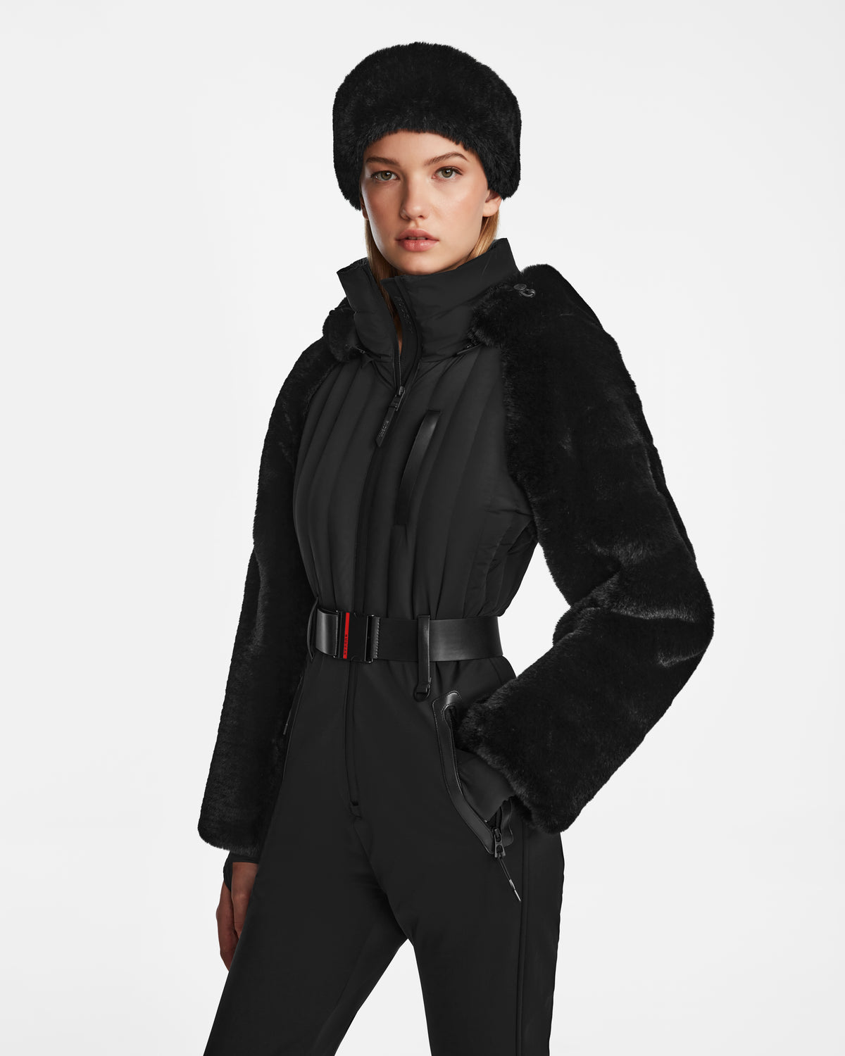 A woman stands against a plain white background, wearing the Rudsak DESTINY - 8124727 Black one-piece ski suit with a belt, faux fur sleeves, and a matching faux fur hat.