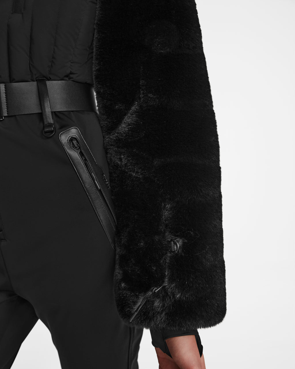 A person wearing Rudsak’s DESTINY - 8124727 Black pants with zippered pockets, a black belt, and a black faux fur coat draped over their arm, styled like a chic women’s ski suit against a plain white background.