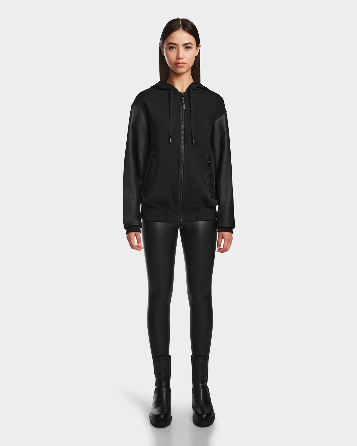 A woman stands facing forward wearing the DRAN - 9524501 Black W zip-up hoodie by Rudsak with leather sleeves, paired with black faux leather leggings and ankle boots against a plain white background.
