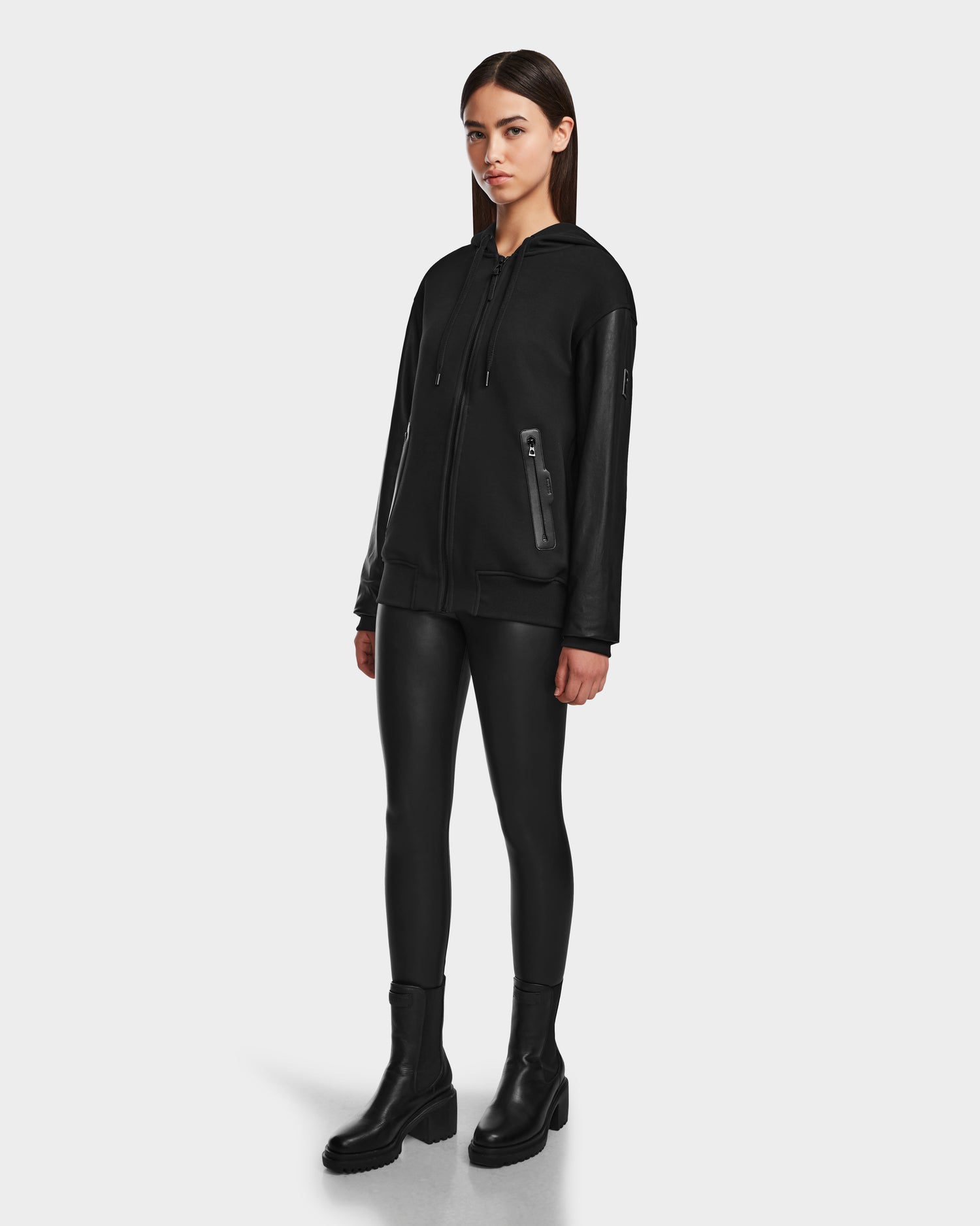 A woman faces forward against a white background, wearing the Rudsak DRAN - 9524501 Black W unisex zip-up hoodie with leather sleeves, paired with black faux leather leggings and ankle boots.