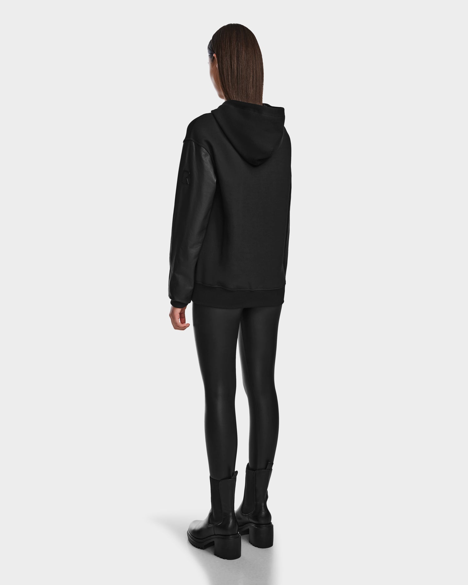 A person with straight brown hair wears the Rudsak DRAN - 9524501 Black W unisex zip-up hoodie with leather sleeves, black leggings, and black boots, standing facing away against a plain white background.