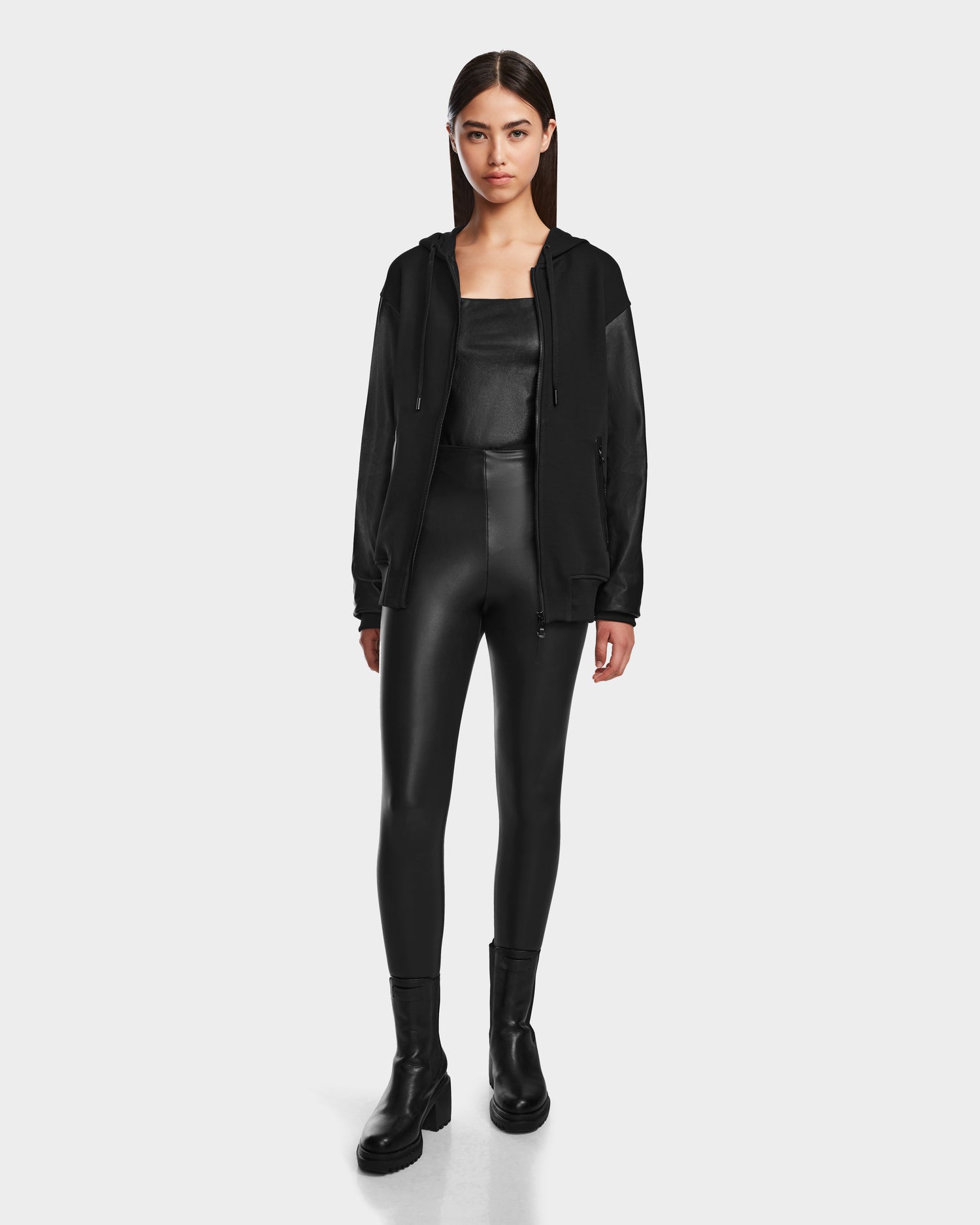 A woman faces forward against a light gray background, wearing the Rudsak DRAN - 9524501 Black W unisex zip-up hoodie with a black top, black high-waisted leather pants, and black boots.