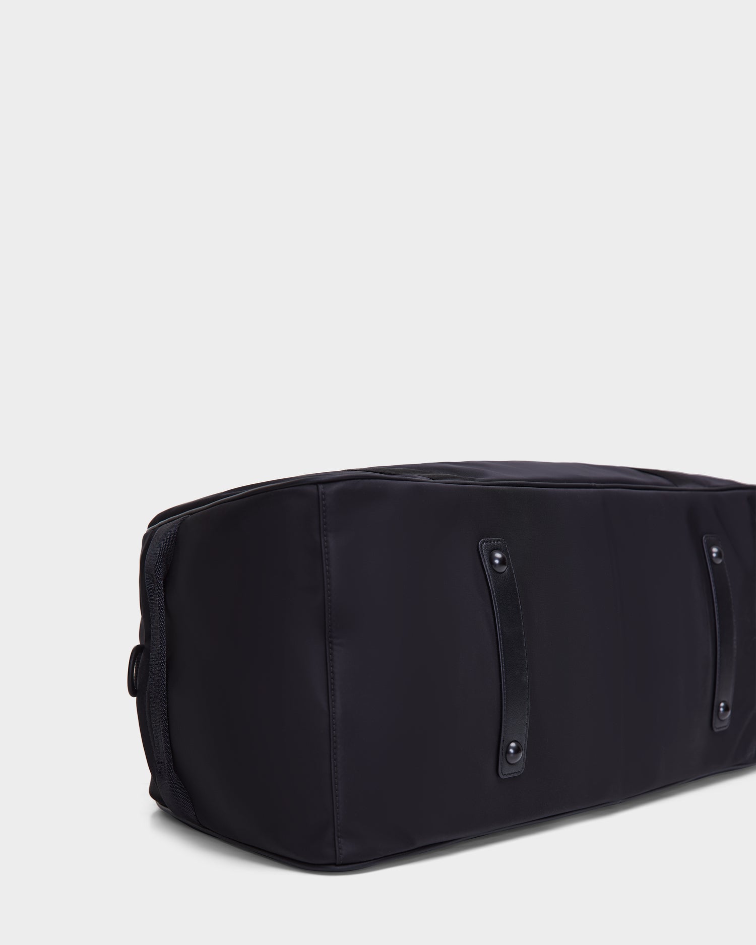 A close-up of the bottom of the Rudsak DUFF - 9324506 Black unisex duffel bag shows two parallel protective strips and four metal rivets against a light background.