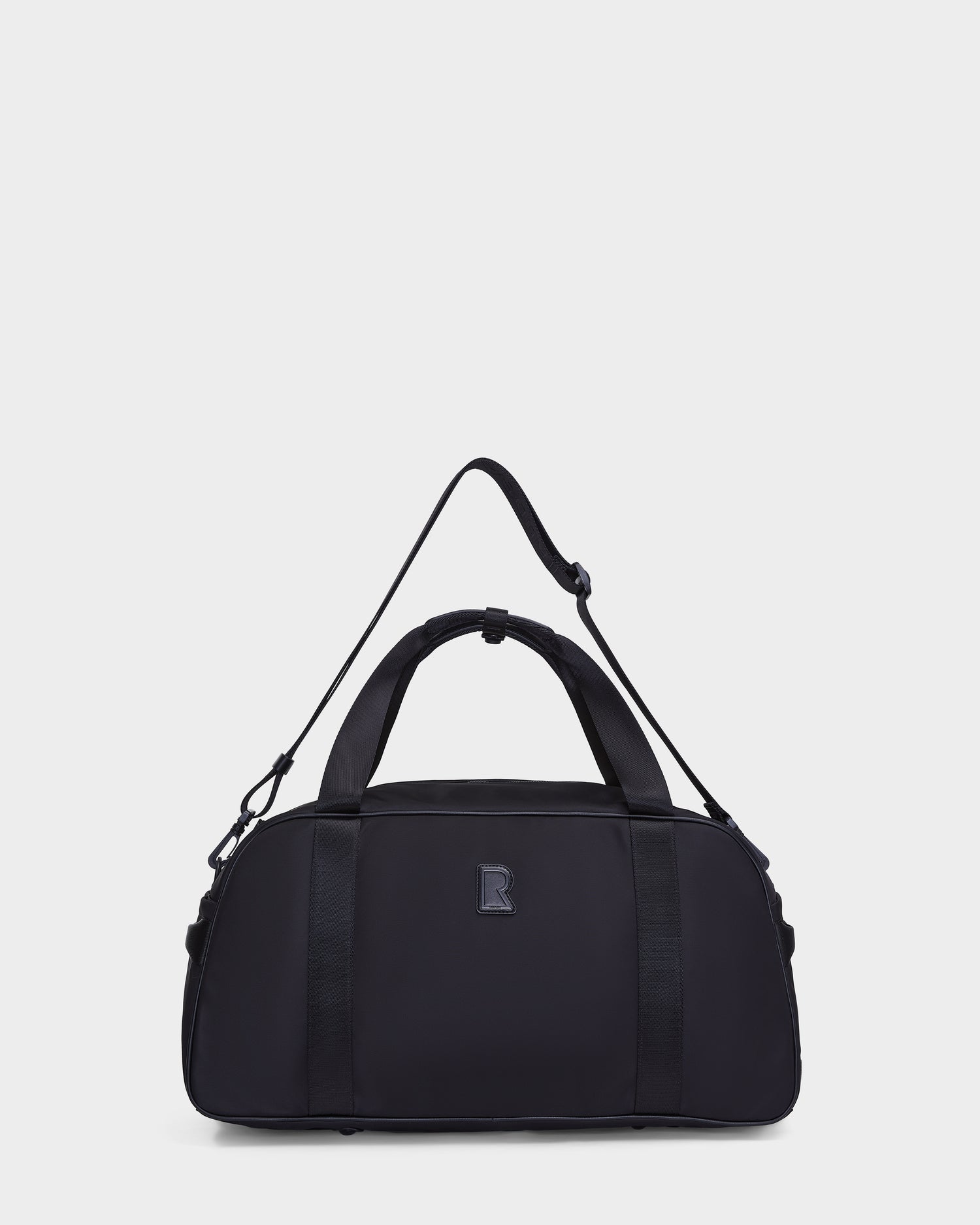 The Rudsak DUFF - 9324506 Black is a black unisex duffel bag with dual handles, an adjustable shoulder strap, and a subtle R logo on the front, shown against a plain light gray background.