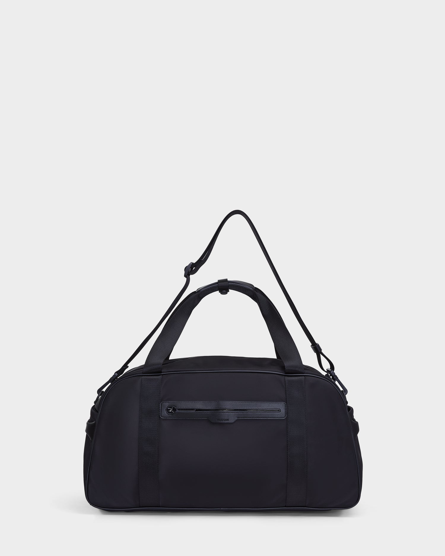 The Rudsak DUFF - 9324506 Black is a unisex duffel bag with two short handles, an adjustable shoulder strap, and a front zip pocket, shown on a plain white background.