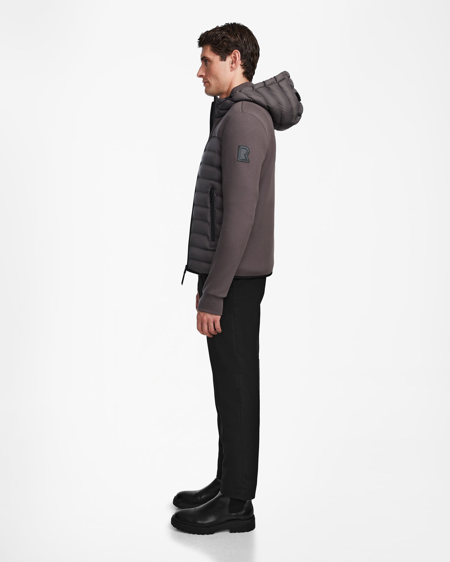 A man stands in profile against a white background, wearing Rudsaks DURIN M - M123515 Grey men’s hooded jacket with lightweight down insulation, paired with black pants and black boots.