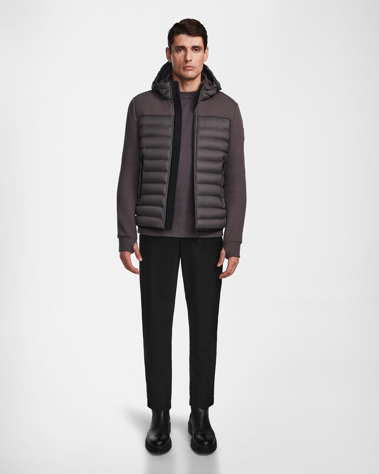 A man stands facing forward, wearing the Rudsak DURIN M - M123515 Grey mens hooded jacket with lightweight down insulation, paired with black pants and black Chelsea boots against a plain white background.