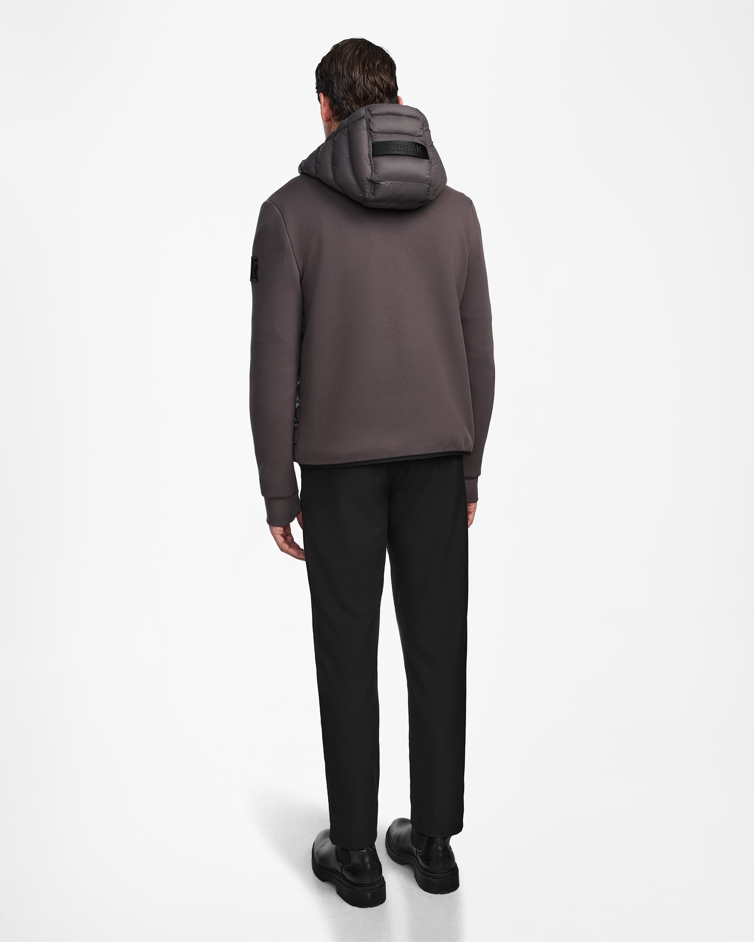 A man, facing away, wears the Rudsak DURIN M - M123515 Grey mens hooded jacket with waterproof zippers, black pants, and black shoes against a plain white background.