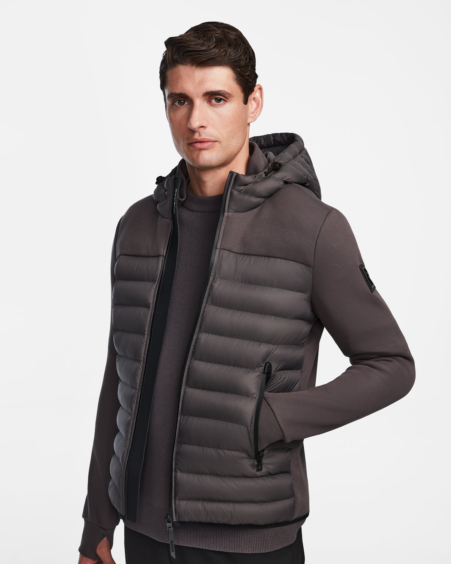 A man with short brown hair wears the Rudsak DURIN M - M123515 Grey men’s hooded jacket with lightweight down insulation and waterproof zippers, layered over a matching sweater, standing against a plain white background.
