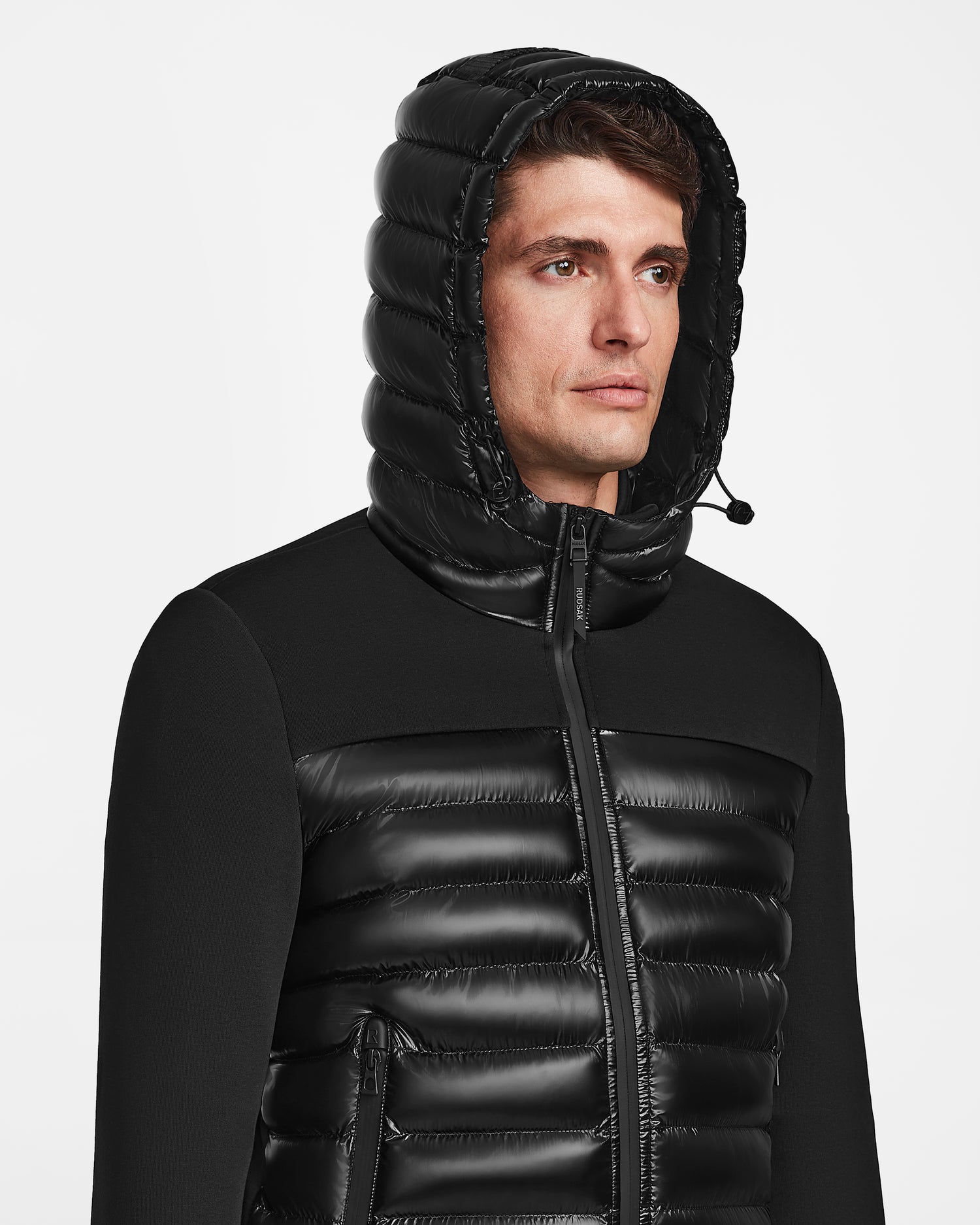 A man models the Rudsak DURIN S - M123518 Black mens hooded jacket, featuring lightweight down insulation and a zip-up front, shown from the chest up against a plain white background.