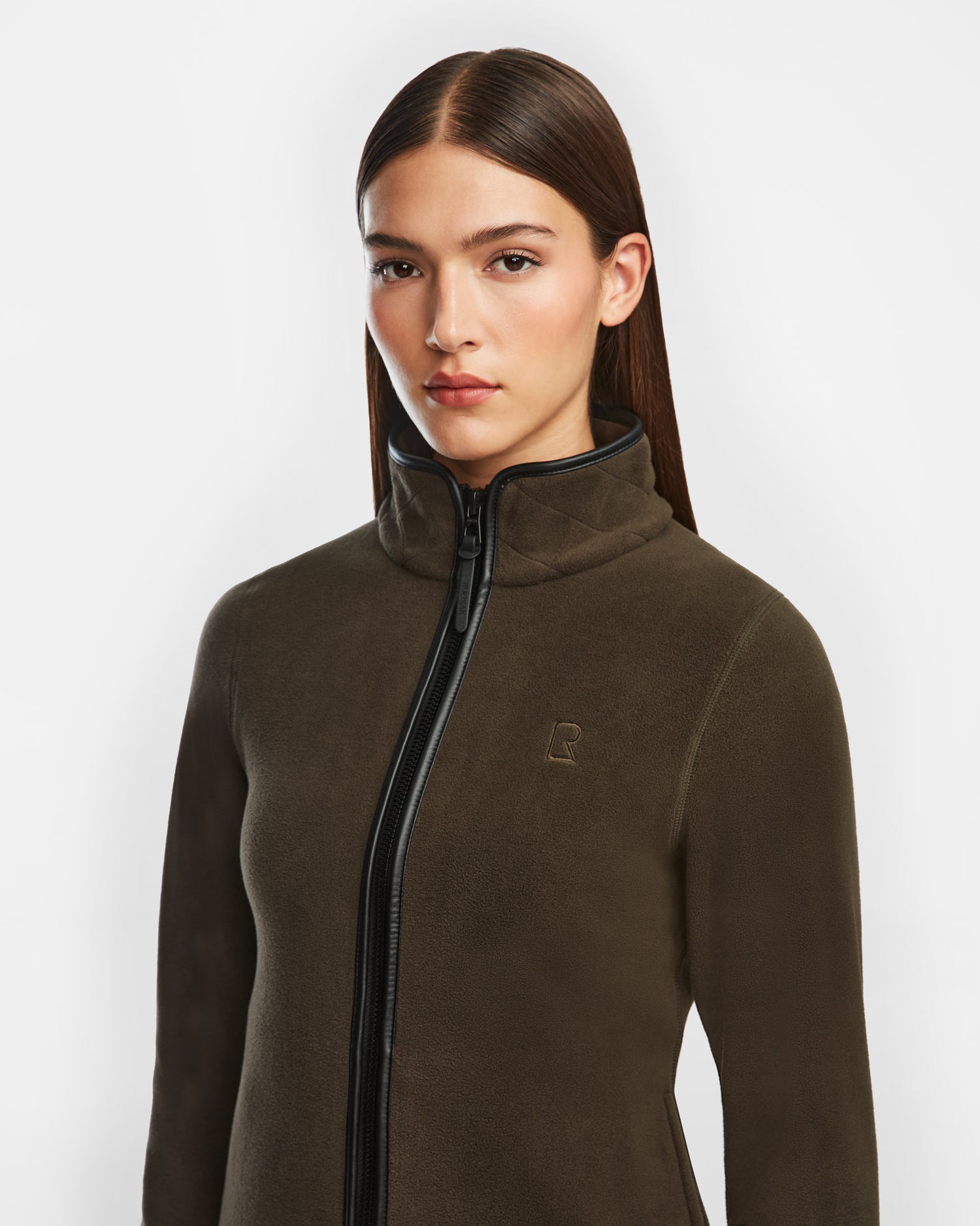 A woman with straight brown hair wears the Rudsak EDITH - 8524617 Army zip-up sweater in dark olive, featuring a high collar, subtle chest logo, soft polar fleece, and vegan leather trim, standing against a plain white background.