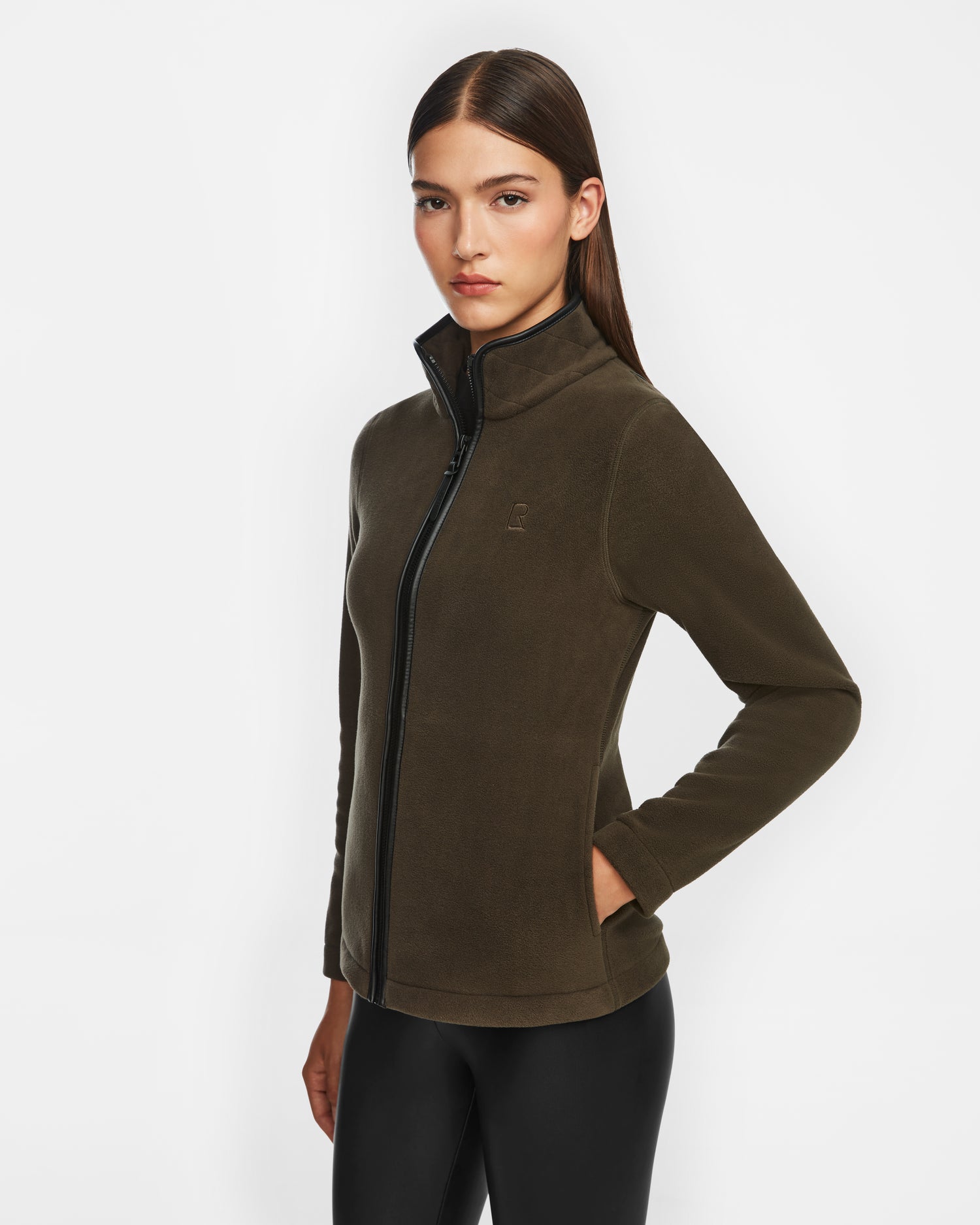 A woman with straight brown hair wears the Rudsak EDITH - 8524617 Army zip-up sweater, styled with black leggings, standing against a plain white background.