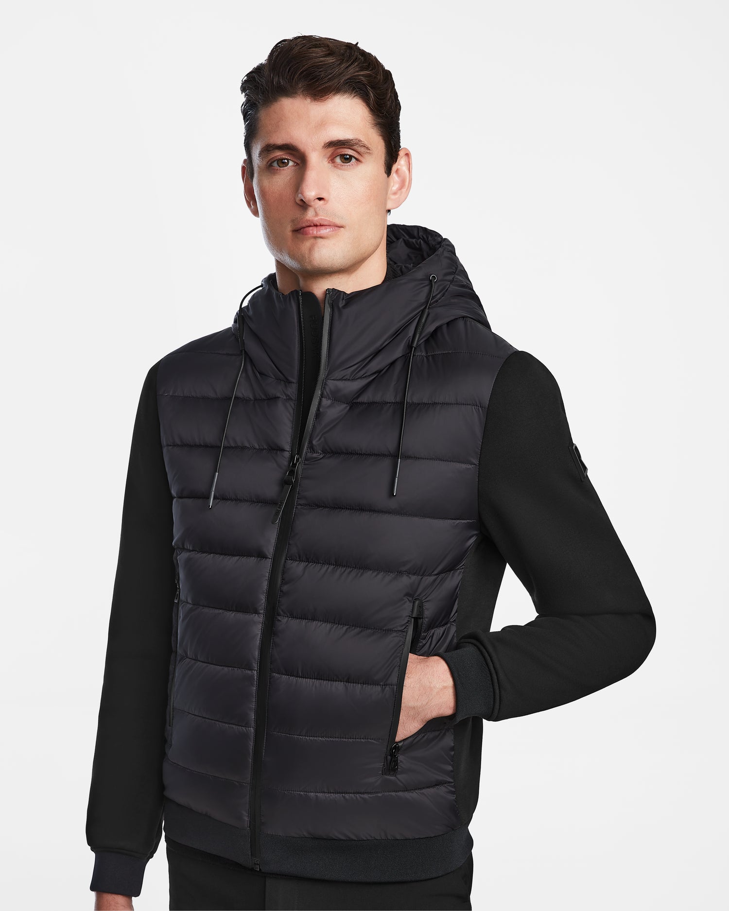 A man stands facing the camera in a Rudsak ELIAS - M124002 Black mens quilted light jacket with a hood and zippered pockets, set against a plain white background.