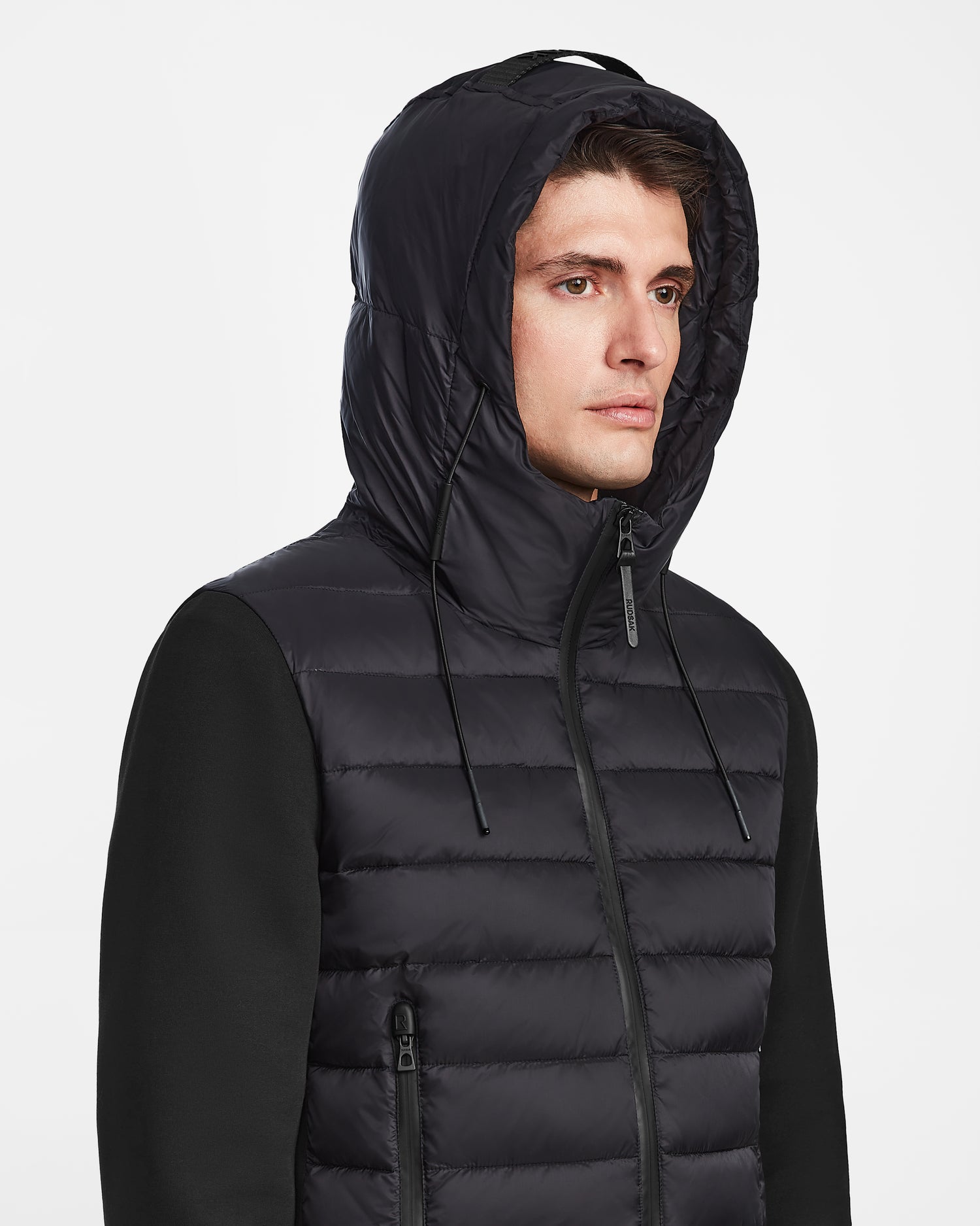 A young man wearing the Rudsak ELIAS - M124002 Black men’s quilted light jacket with hood up looks off to the side. The wind- and water-resistant jacket features zipper pockets, hood drawstrings, and is set against a plain white background.