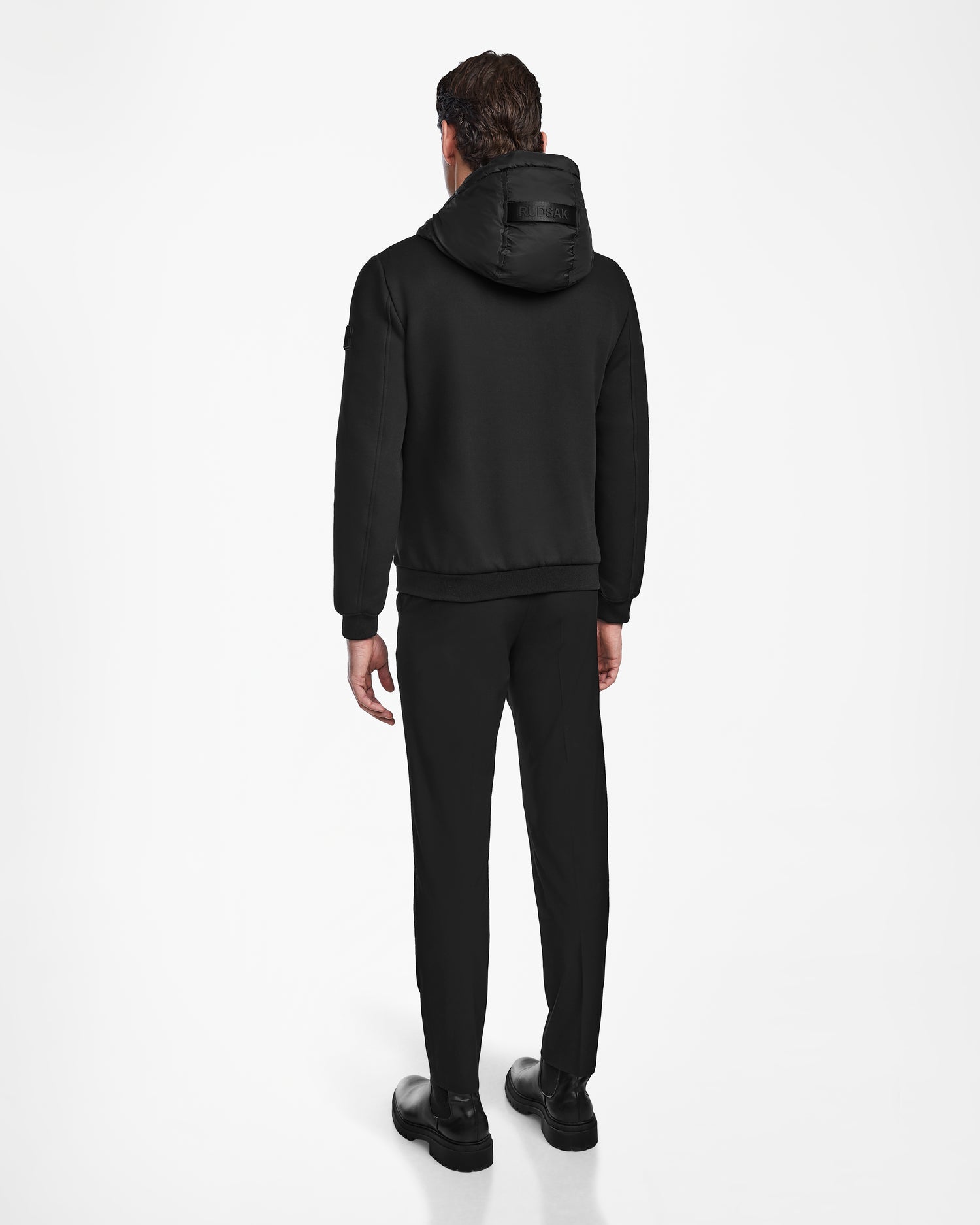 A man faces away from the camera, wearing the Rudsak ELIAS - M124002 Black quilted mens jacket, paired with black pants and shoes against a plain white background.