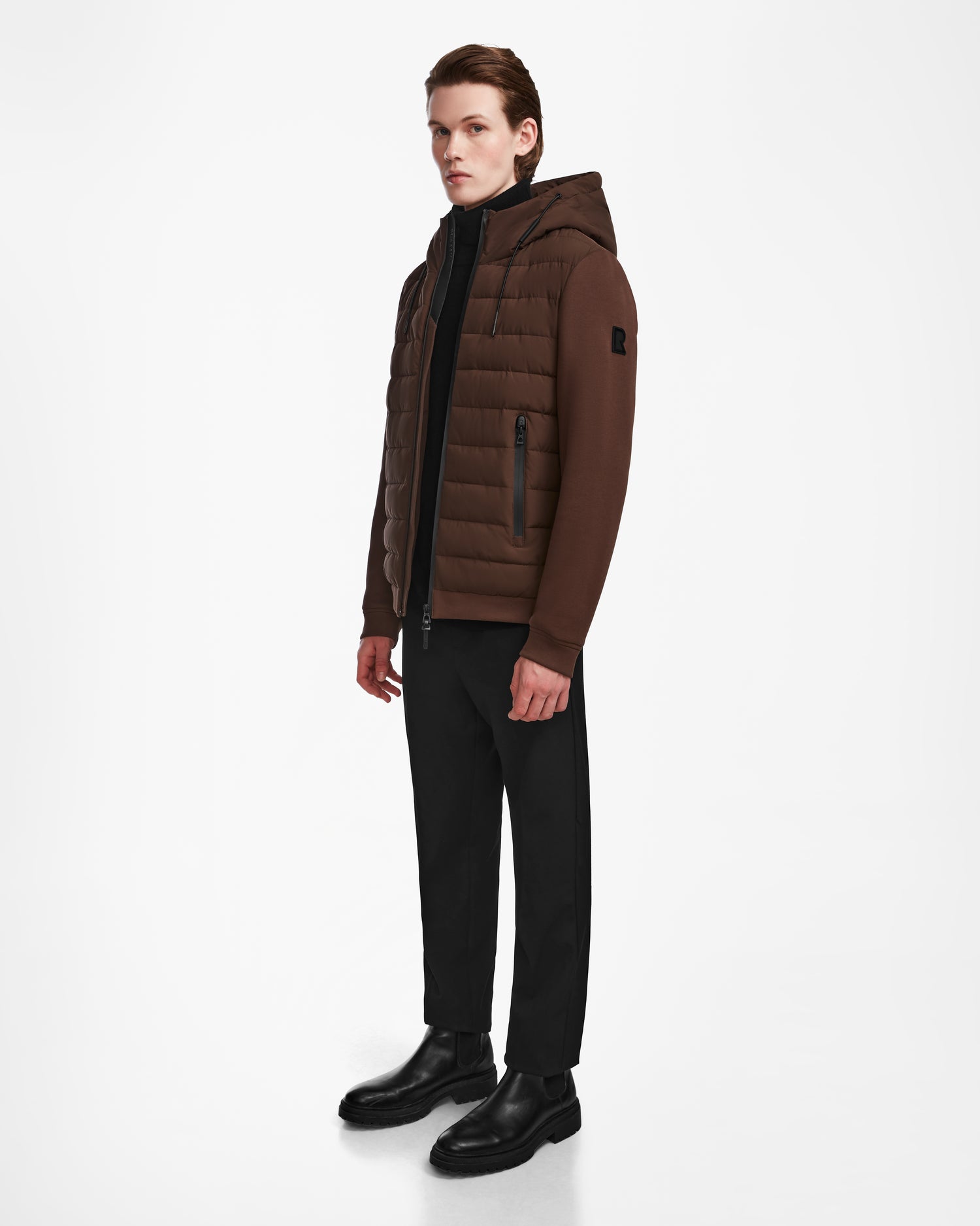 A young man stands against a white background wearing the Rudsak ELIAS - M124002 Espresso men’s quilted jacket with ecofill, black pants, and black boots. He is facing slightly to the side with a neutral expression.
