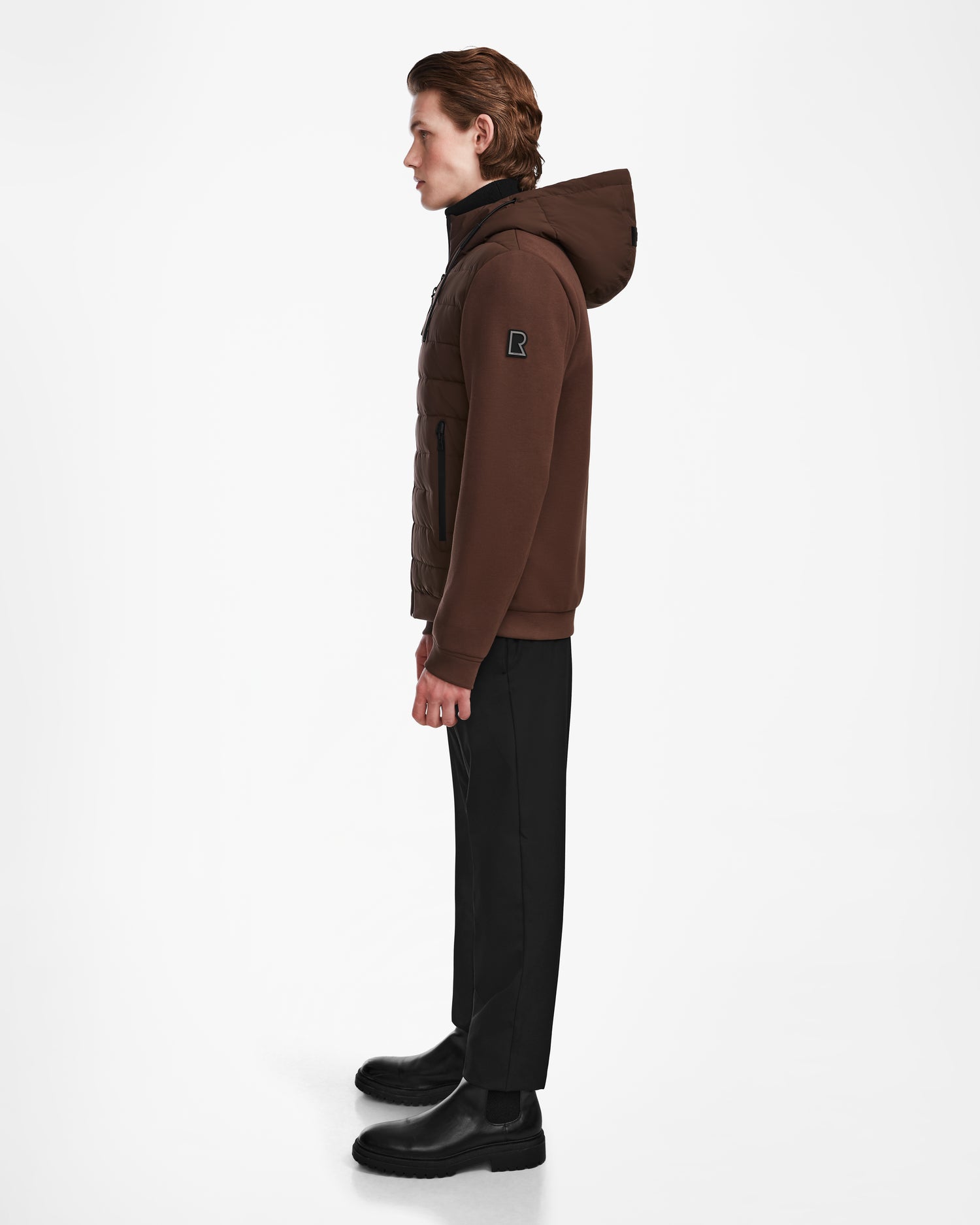 A person with brown hair stands in profile against a white background, wearing the Rudsak ELIAS - M124002 Espresso mens quilted light jacket, black pants, and black boots.