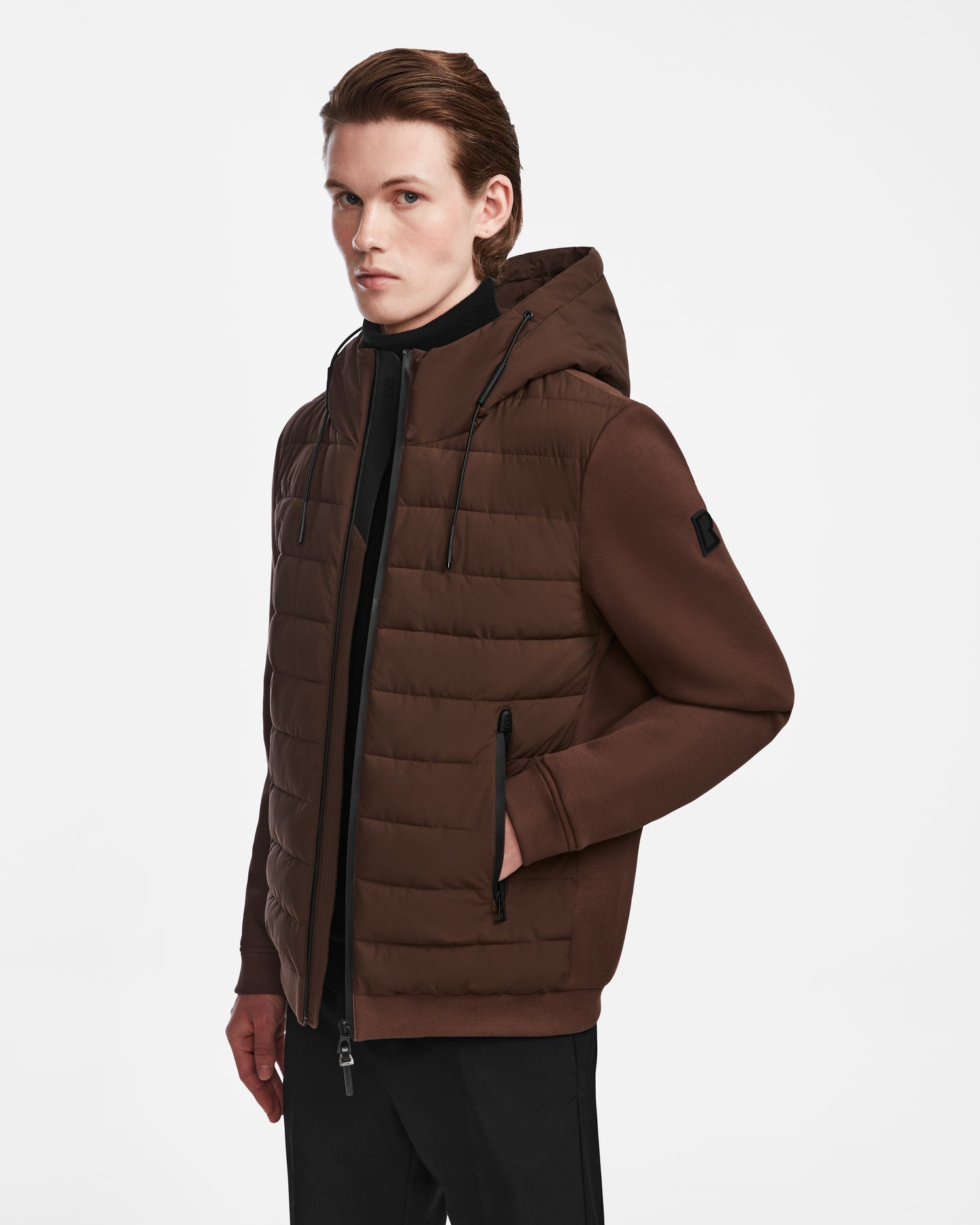 A person with light skin and brown hair wears the Rudsak ELIAS - M124002 Espresso men’s quilted jacket and black pants, standing with one hand in their pocket against a plain white background.