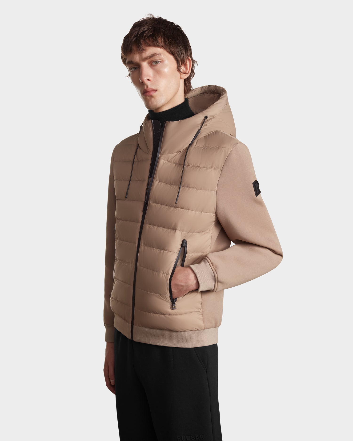 A young man poses with one hand in his pocket wearing the Rudsak ELIAS - M124002 Latte men’s quilted light jacket, featuring wind- and water-resistant fabric, a front zipper, and black drawstrings, against a plain white background.