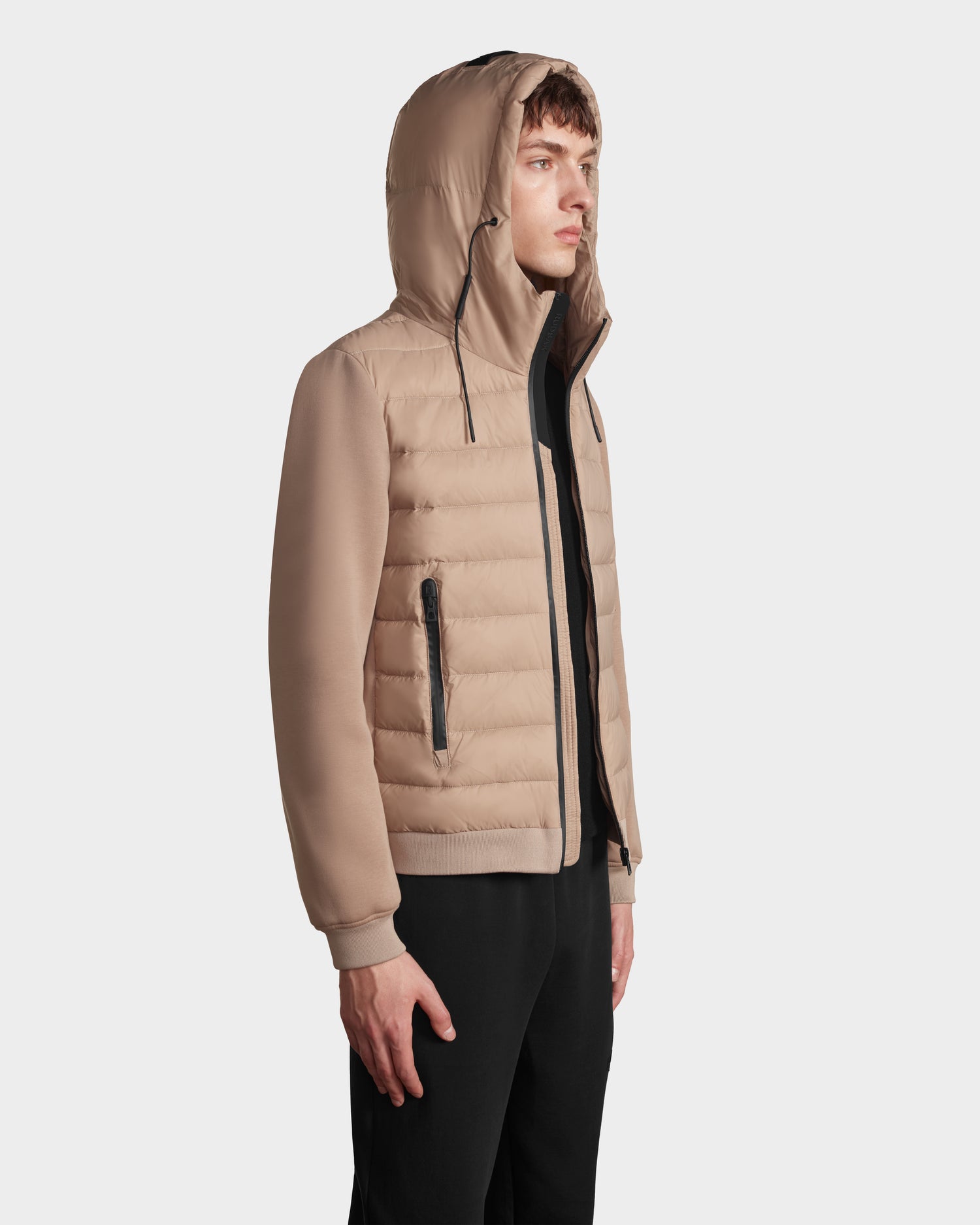 A young man stands in profile, wearing Rudsaks ELIAS - M124002 Latte quilted light jacket with zippered pockets and black pants, looking forward against a plain light background.